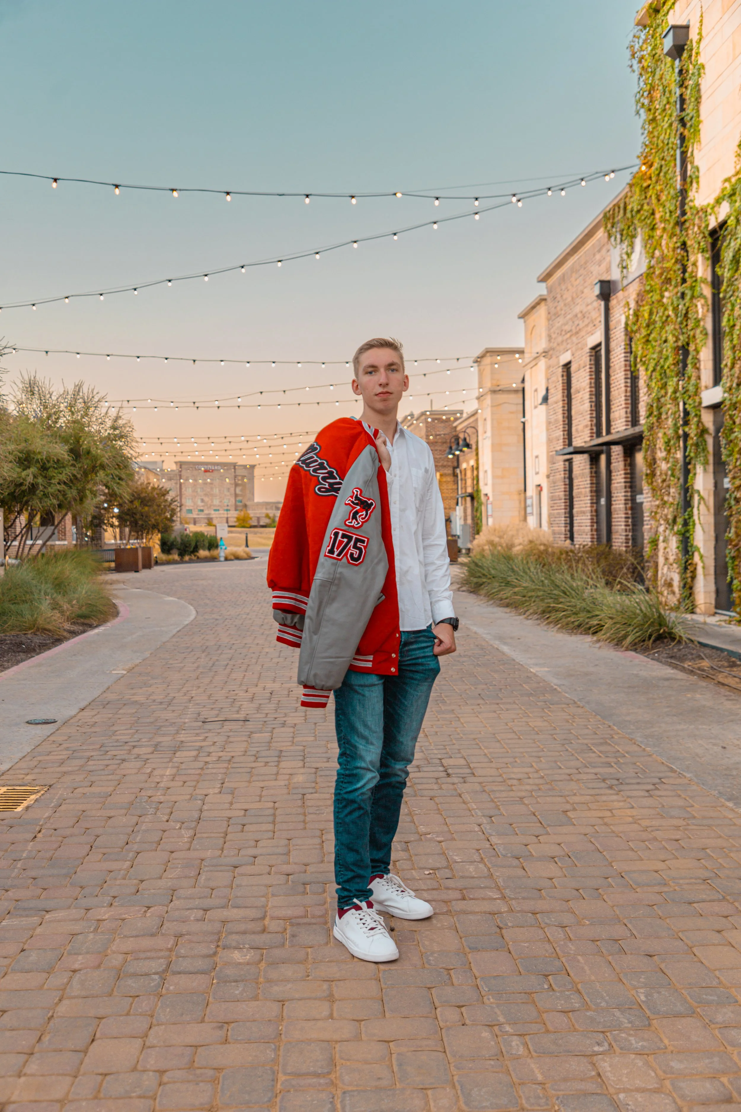 A young man standing on a cobblestone street in an urban area at sunset, wearing a white shirt, blue jeans, white sneakers, and draping a red and gray varsity jacket over his shoulder.