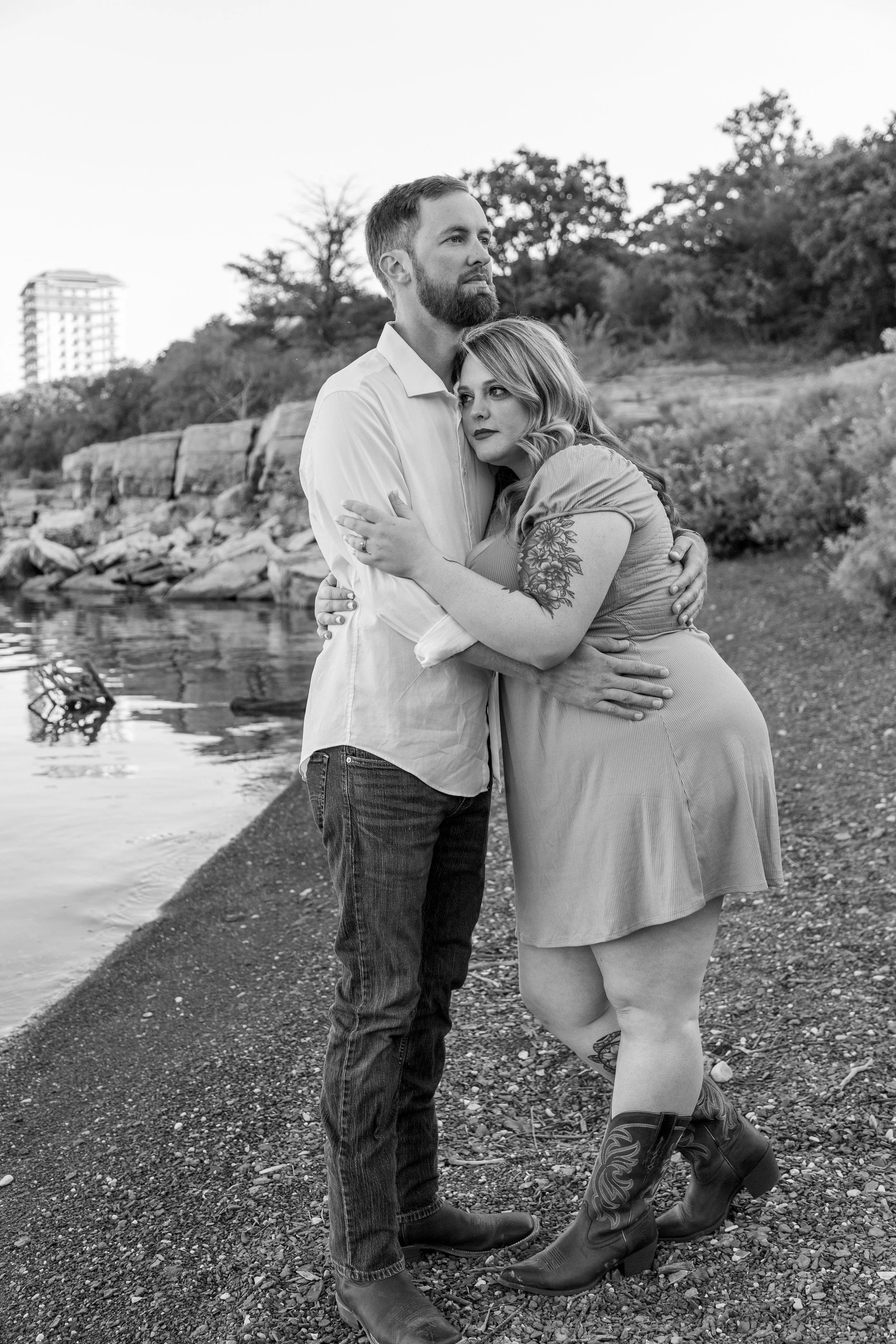A black and white photo of a couple standing by a lakeshore, embracing each other. The man has a beard and wears a light-colored shirt and jeans. The woman has wavy hair, a tattoo on her arm, and wears a short dress and cowboy boots. Trees and rocks 