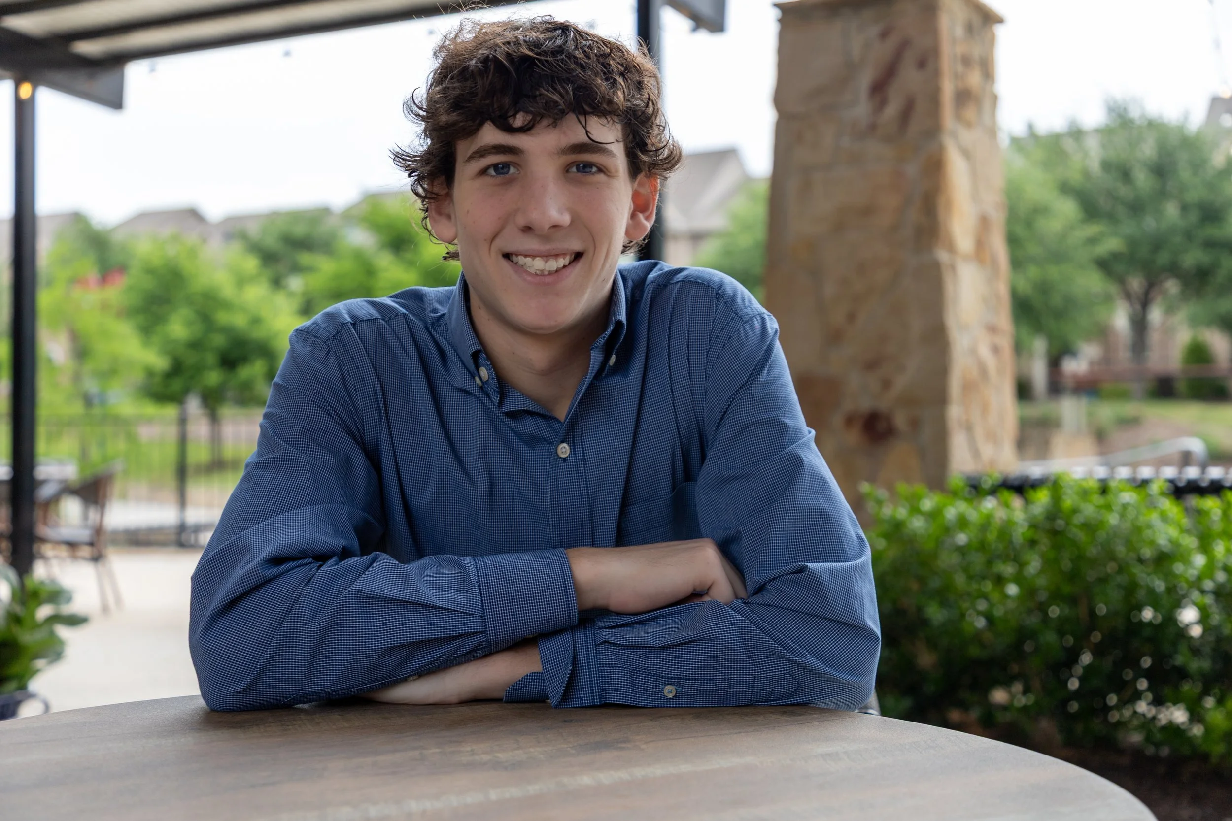 A young man with curly brown hair smiling, sitting at a wooden table outdoors with green trees and a stone pillar in the background.