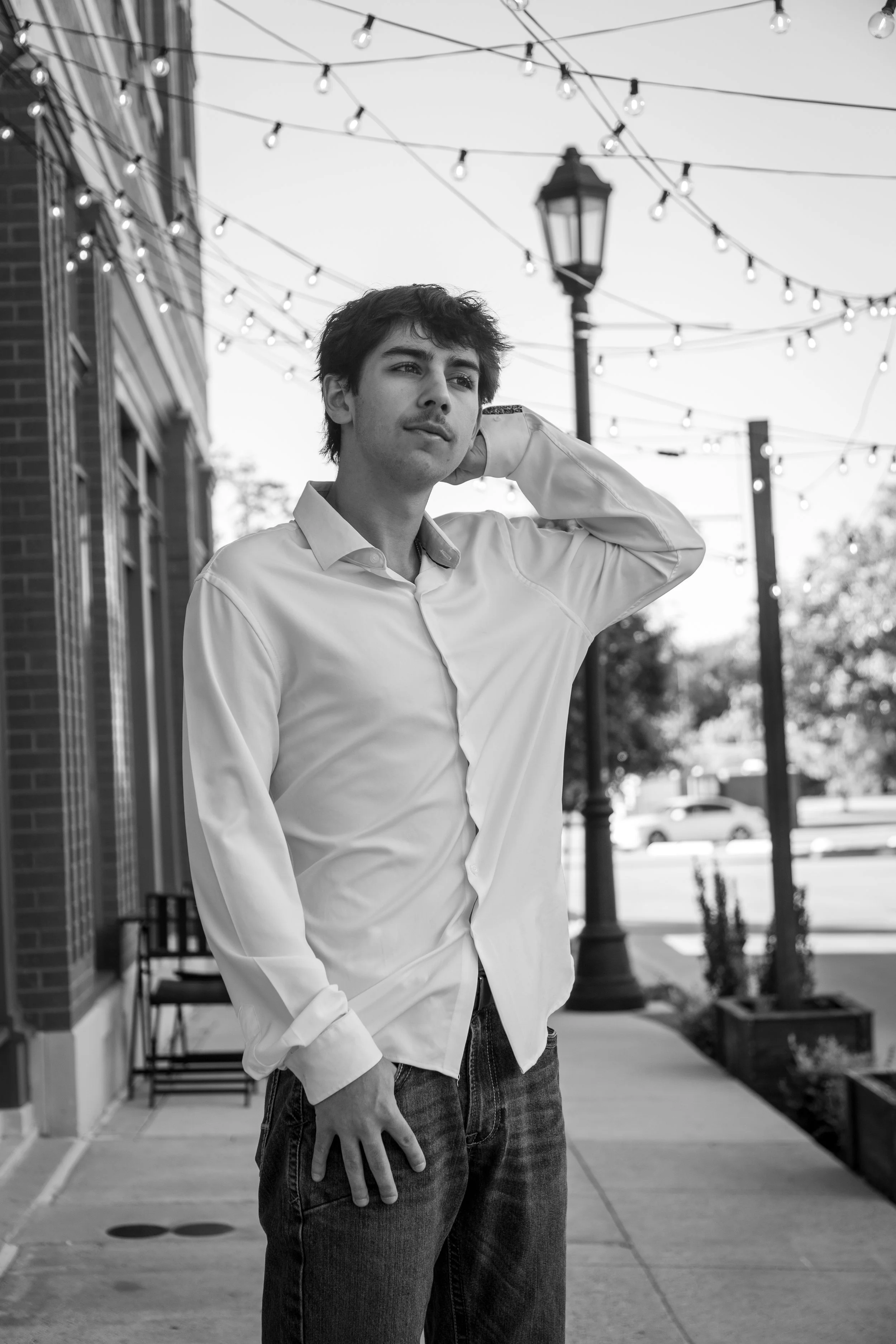 A young man in a white button-up shirt and jeans standing on a sidewalk with string lights overhead and a streetlamp behind him, in an urban outdoor setting.