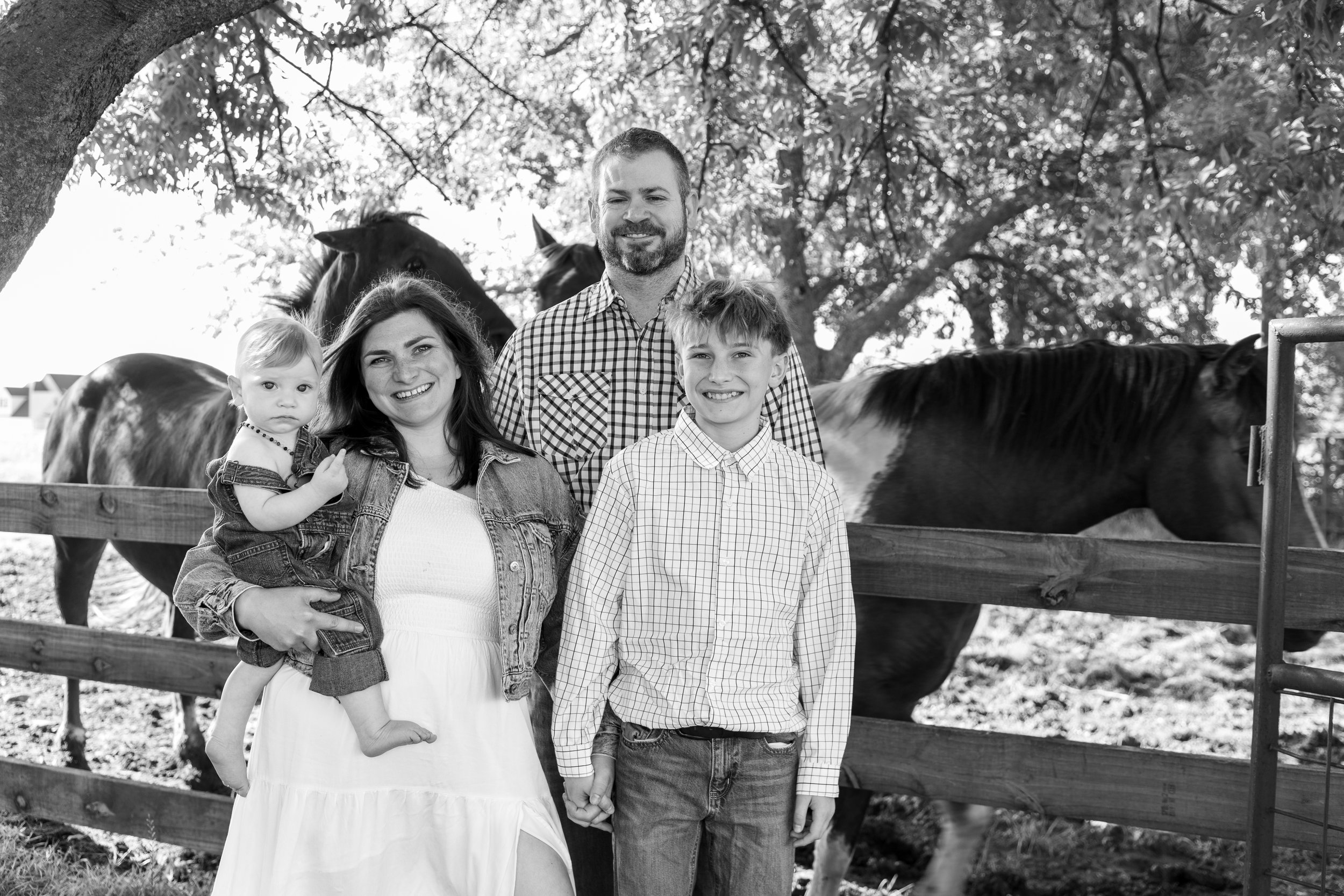 A family of four standing outdoors in front of a wooden fence with horses and trees in the background. The family includes a woman holding a young girl, a man, and a teenage boy.
