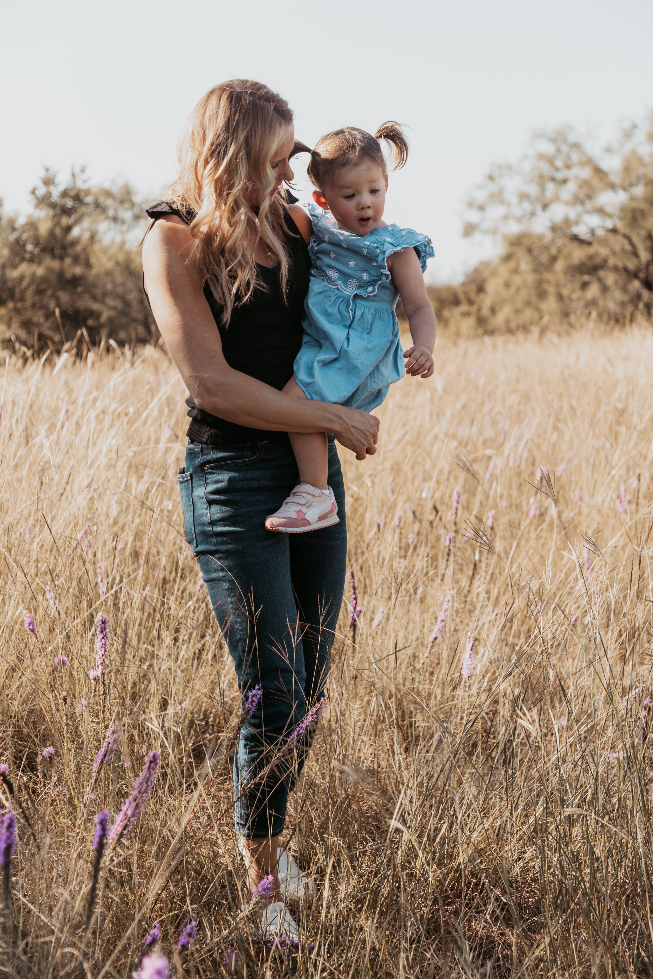 A woman holding a young girl in a field of tall grass and purple wildflowers on a sunny day.
