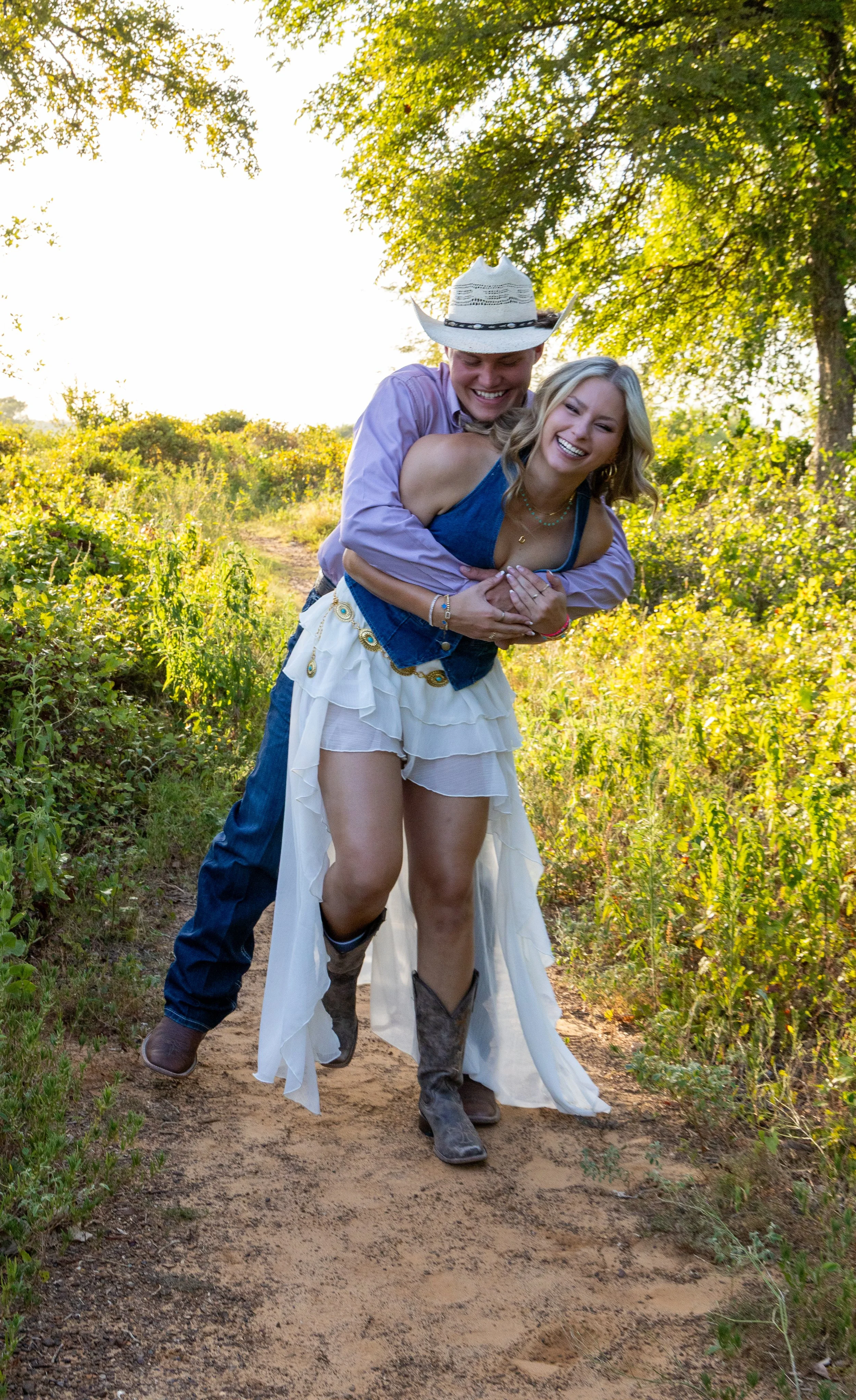 A man and a woman smiling and laughing while walking on a dirt trail in a lush green outdoor setting during daytime. The man is wearing a cowboy hat, a pink shirt, and jeans, while the woman is wearing a blue top, a white layered skirt, and cowboy bo