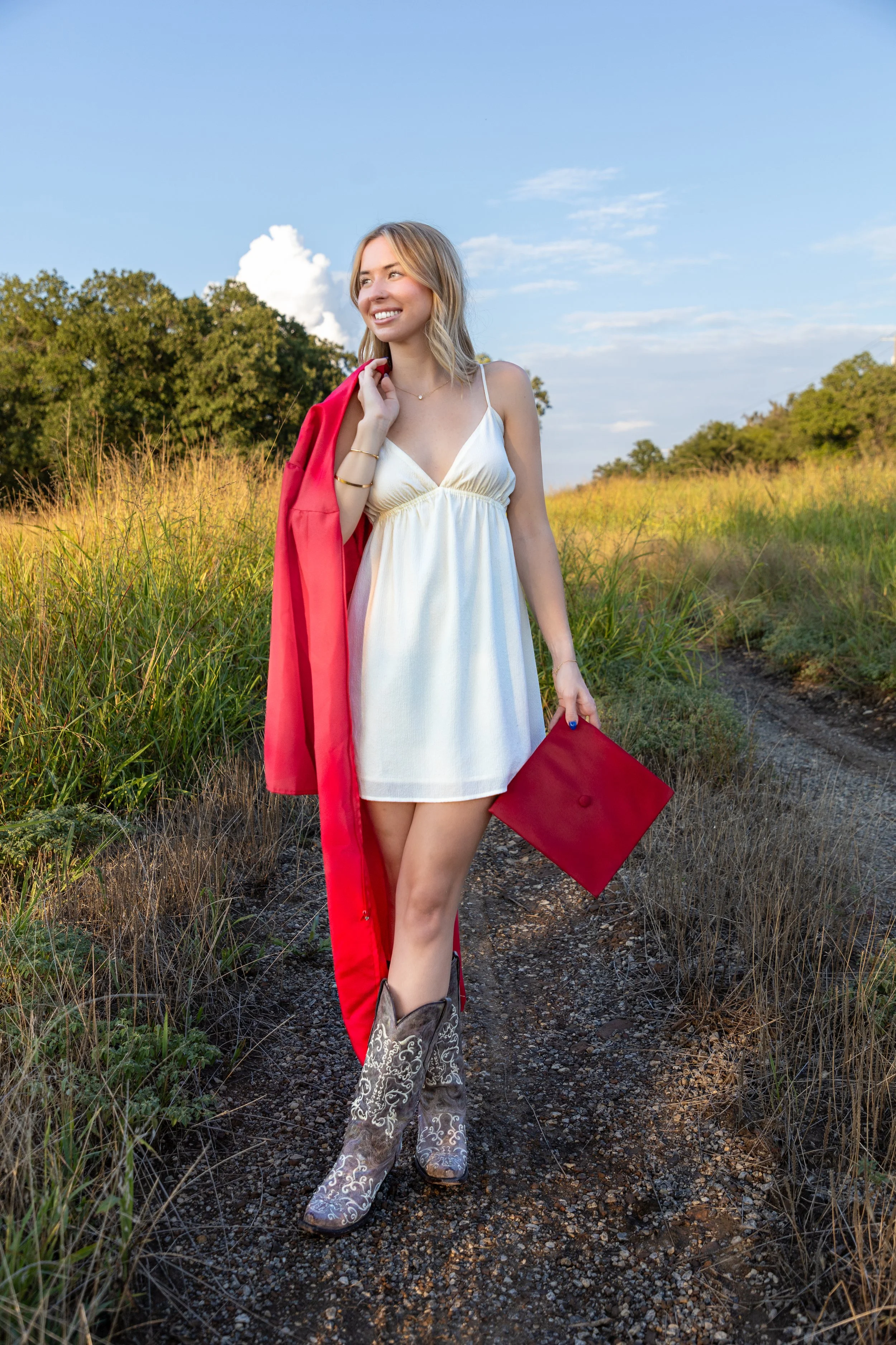 A young woman in a white dress holding a red diploma folder and wearing cowboy boots walks along a dirt trail through a grassy field, carrying a red gown over her shoulder, under a clear blue sky.
