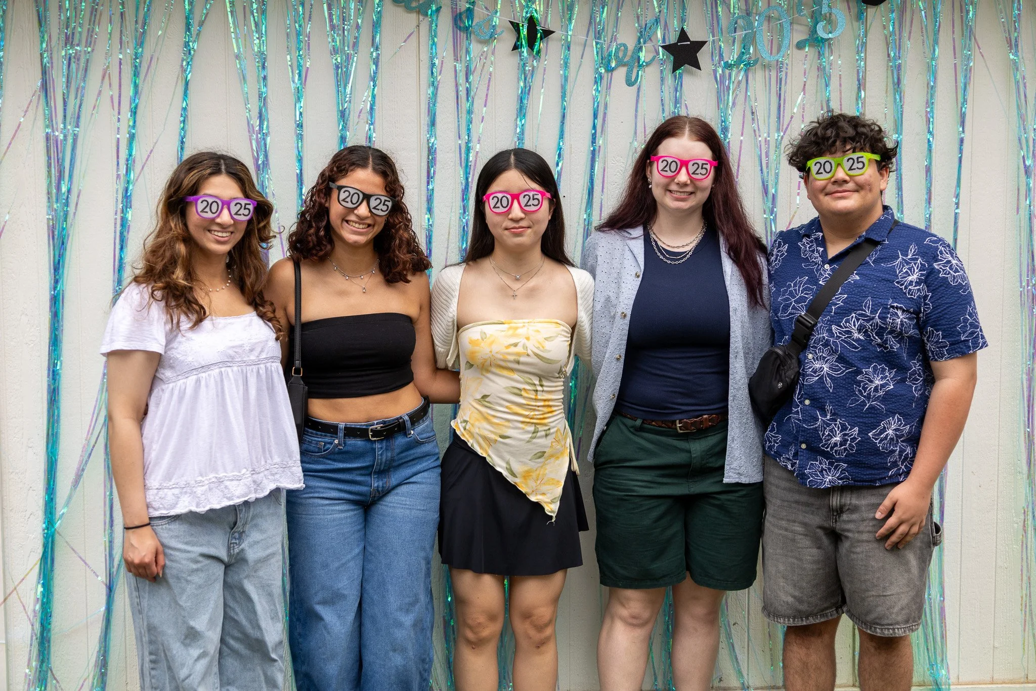 A group of five young people standing in front of a decorative backdrop with tinsel and stars, wearing novelty glasses with age and price tags, smiling and posing for a photo.