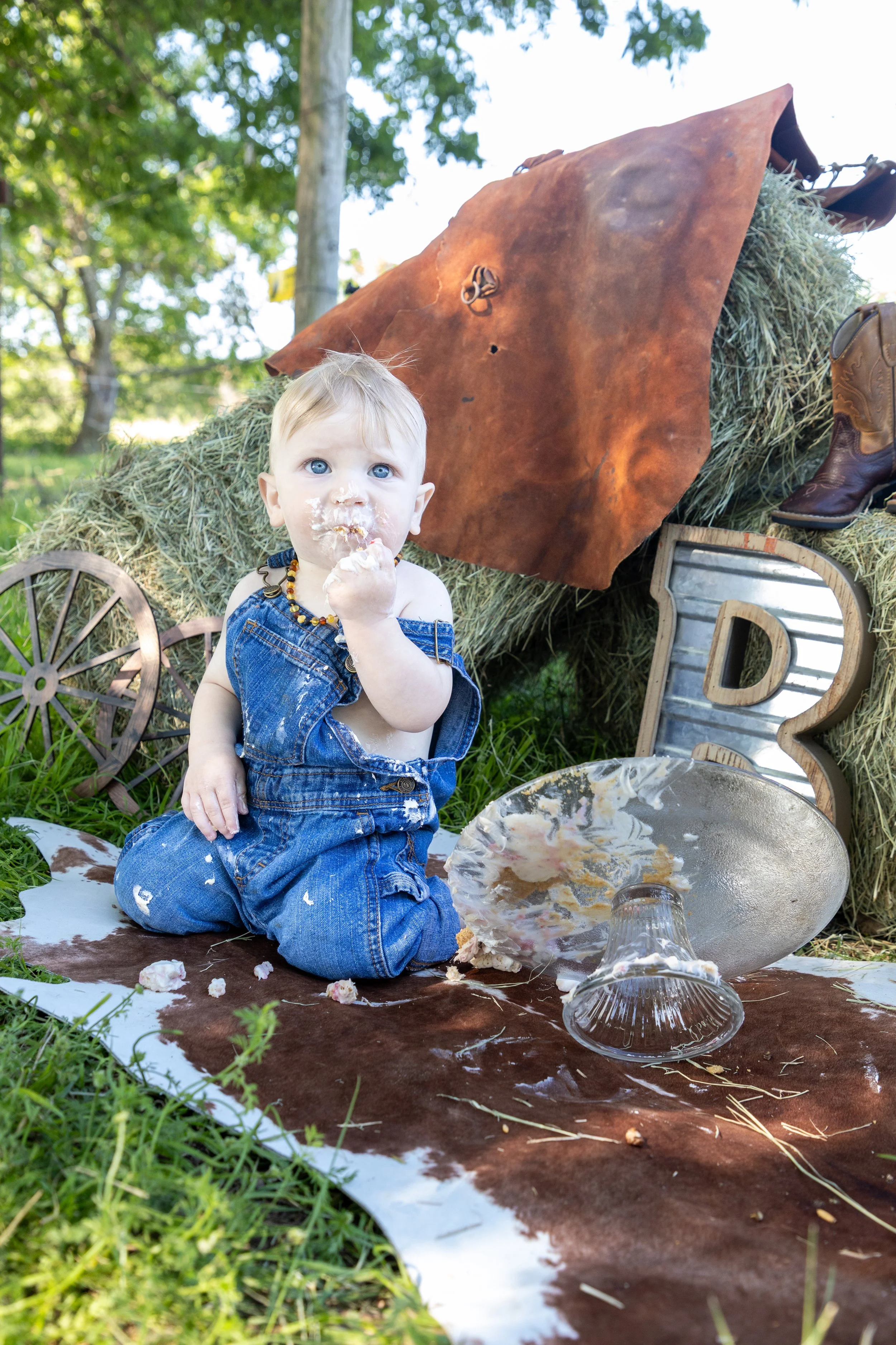 A baby sitting outdoors on a brown and white animal hide, wearing denim overalls, with cake and frosting on their face and hands, surrounded by rustic farm-themed decorations including hay bales, a metal letter 'D', a cowhide rug, a glass bowl, a cow