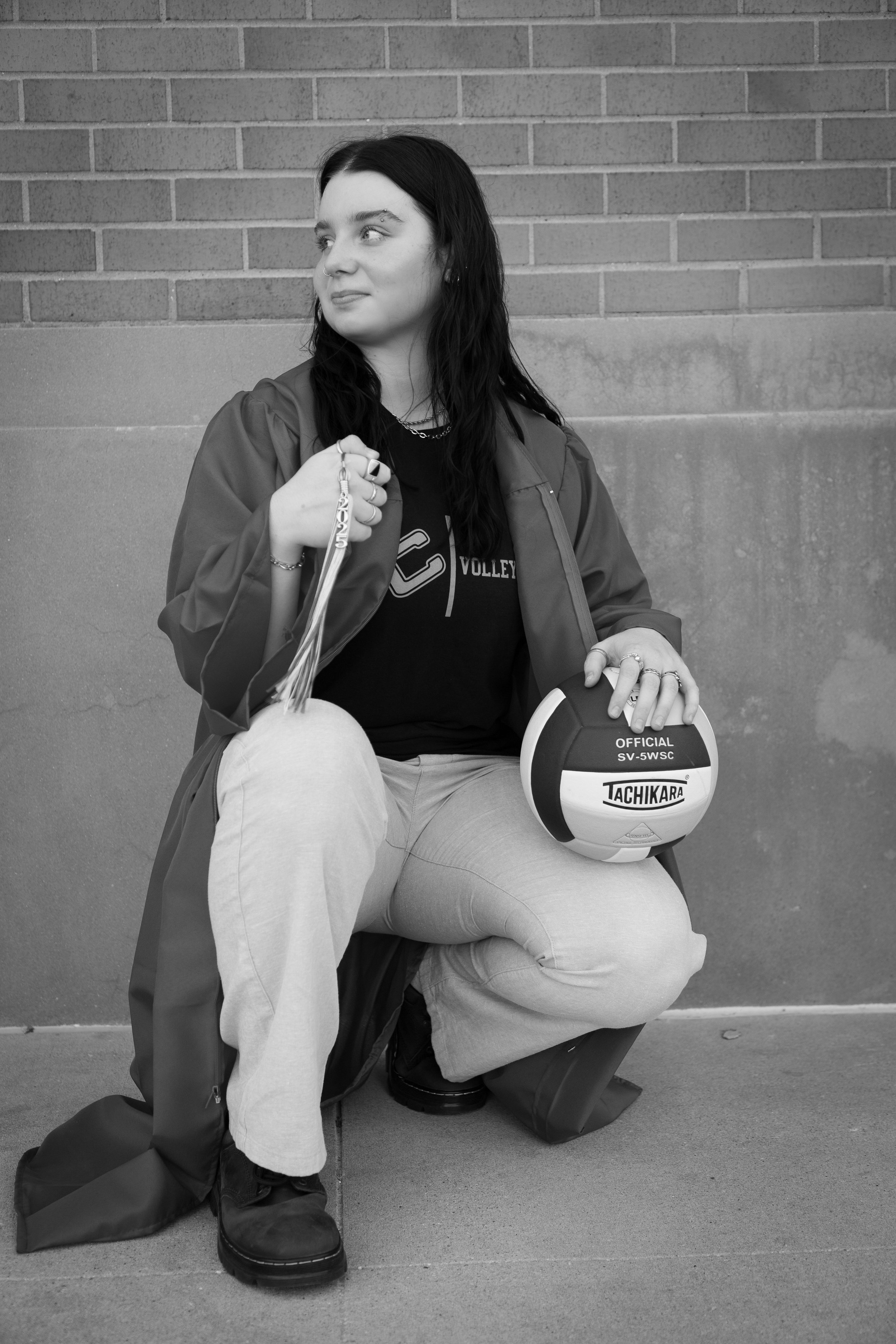 A young woman with long dark hair kneeling on one knee against a brick wall, holding a volleyball in her right hand, wearing a black t-shirt, light-colored pants, and a jacket.