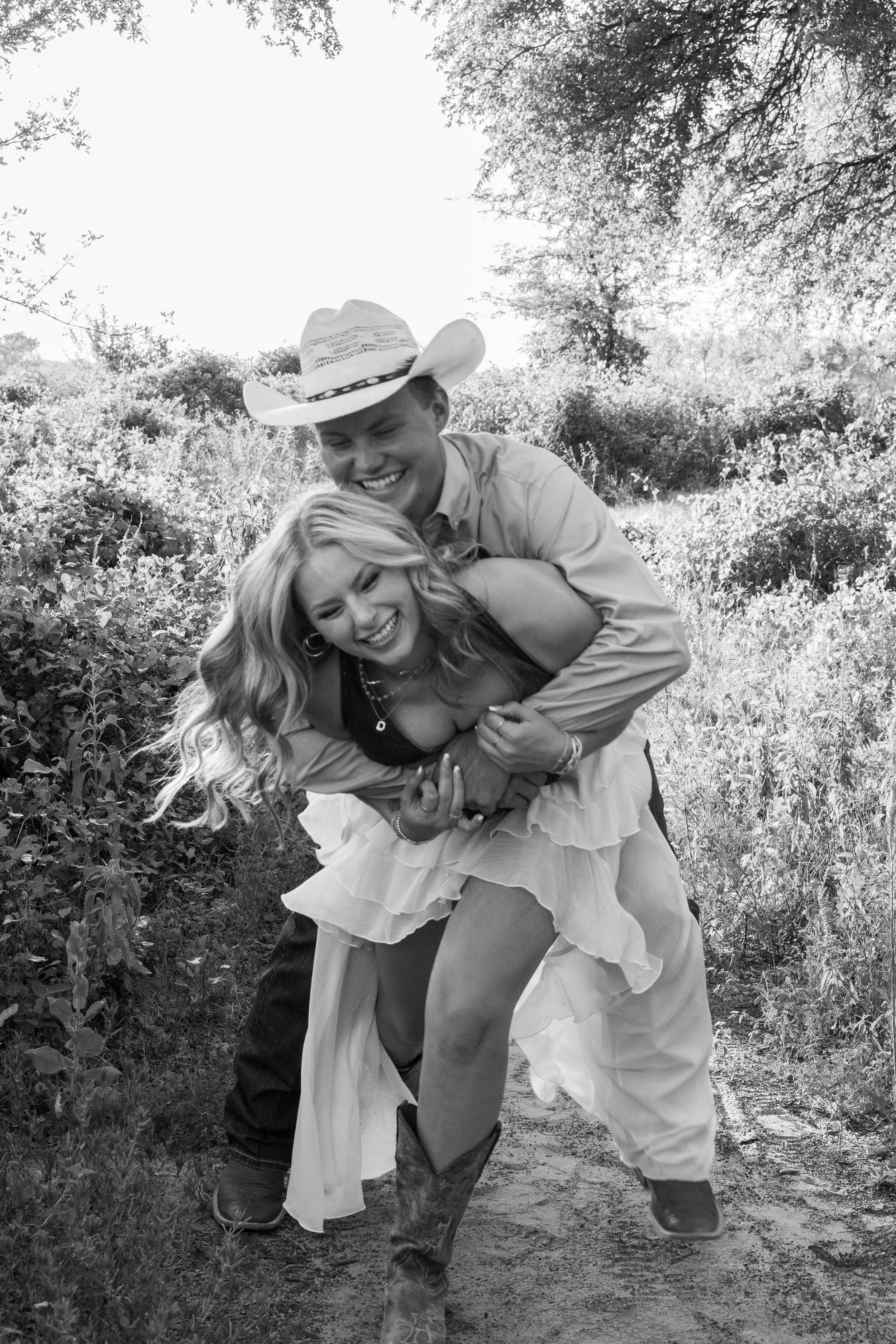 A black and white photograph of a young man and woman outdoors, with trees and bushes in the background. The man, wearing a cowboy hat and Western shirt, is smiling as he leans over and hugs the woman from behind. The woman, with long, wavy hair, is 