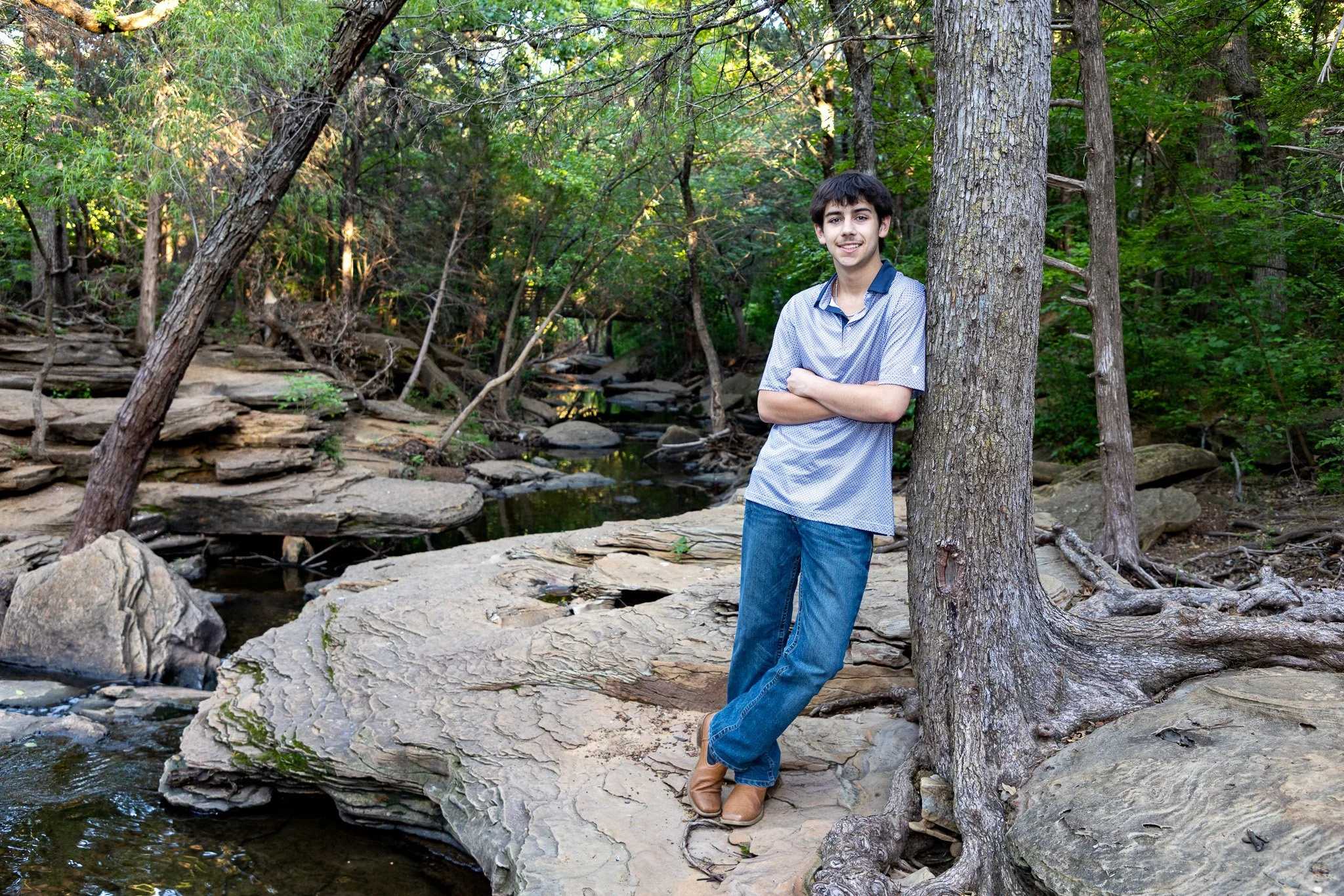 A young man with dark hair is standing outdoors among trees and rocks, leaning against a tree at the edge of a small creek or stream, with arms crossed and smiling.