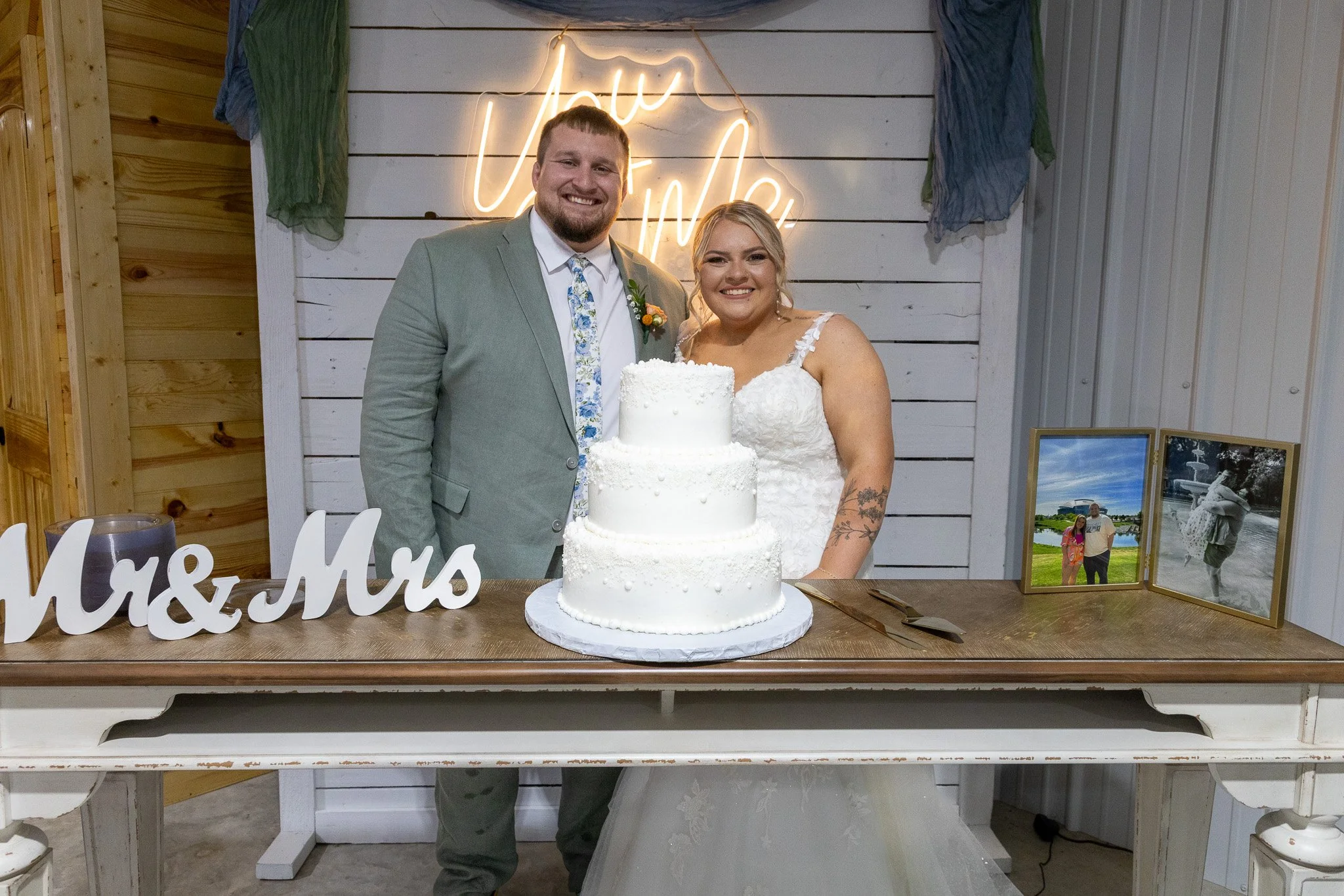 A newlywed couple posing behind a three-tier white wedding cake at their wedding reception. The groom is wearing a light gray suit with a floral tie and boutonniere, and the bride is in a white lace wedding dress with tattooed arms. Behind them is a 