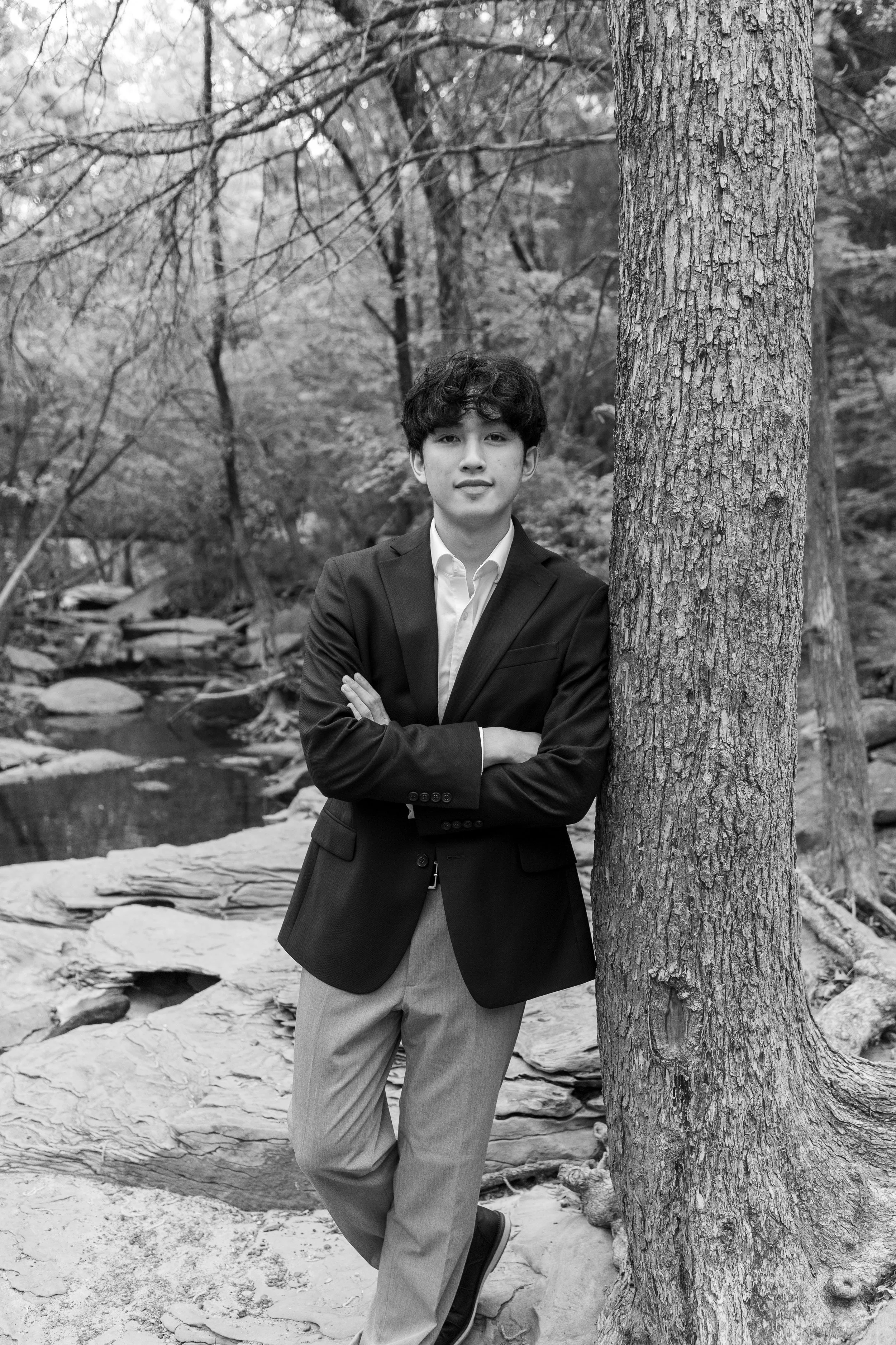 Black and white photo of a young man in a suit standing outdoors next to a tree with crossed arms, with a creek and trees in the background.