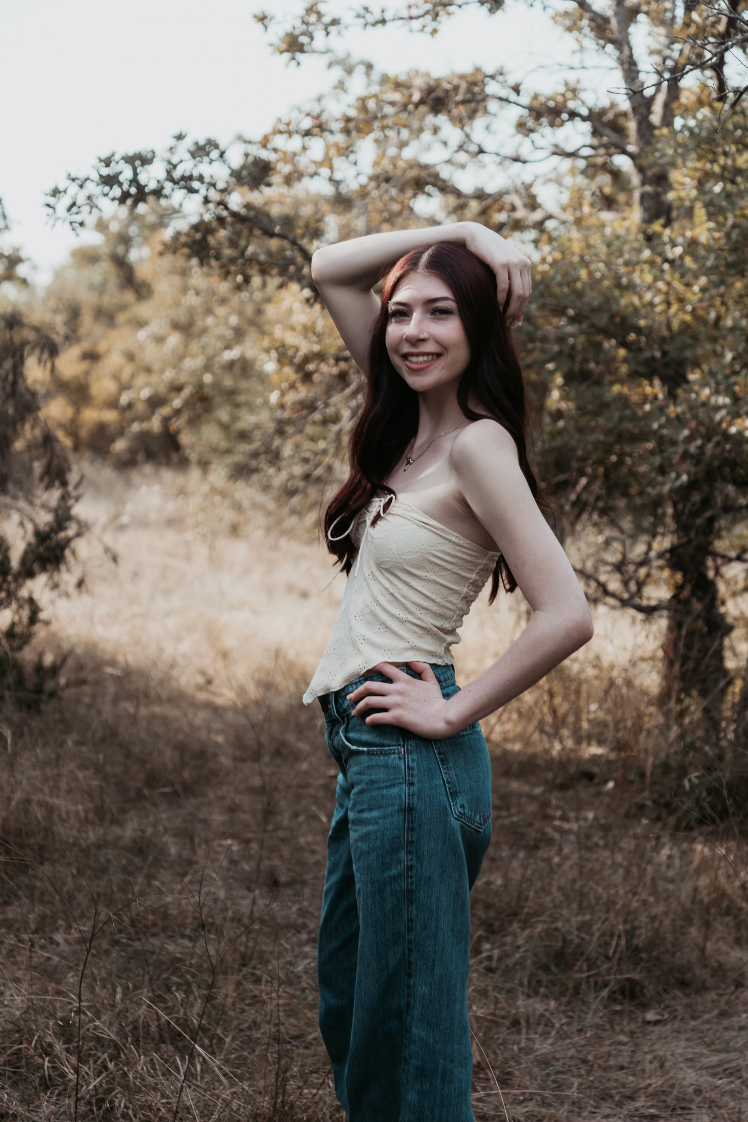 A young woman with long dark red hair, smiling and posing outdoors in a natural setting with trees and dry grass, wearing a light-colored sleeveless top and high-waisted blue jeans.