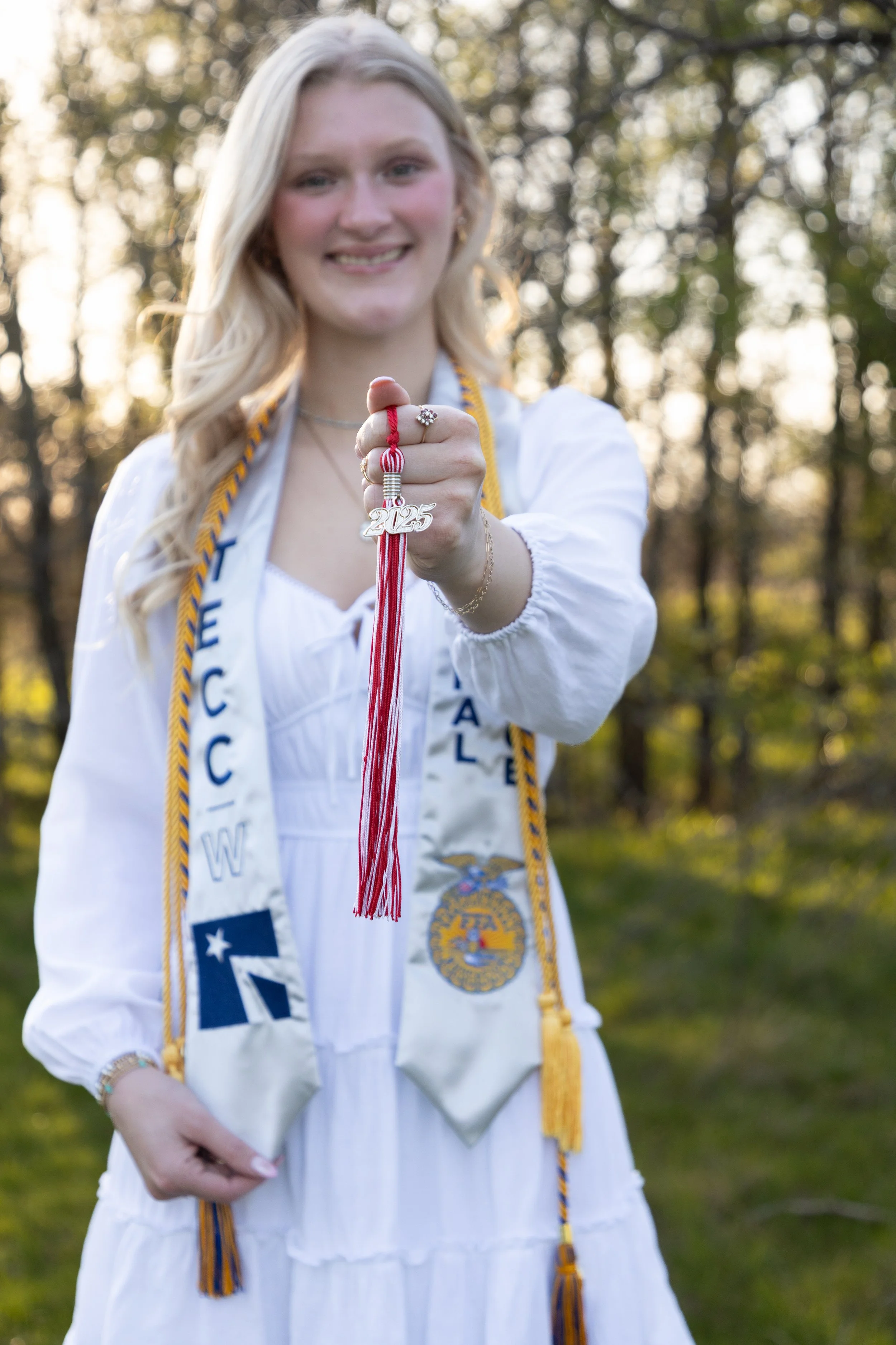 A young woman dressed in graduation attire holding a tassel with the year 2025 on it, outdoors with trees and sunlight in the background.