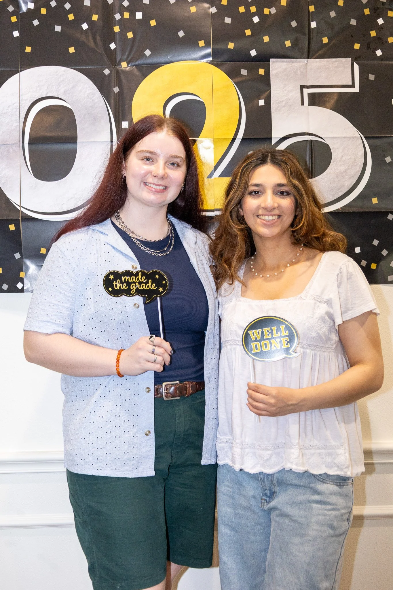 Two young women standing together at a graduation celebration, smiling, with a black and yellow '2023' backdrop behind them. They are holding signs that say 'made the grade' and 'WELL DONE.'