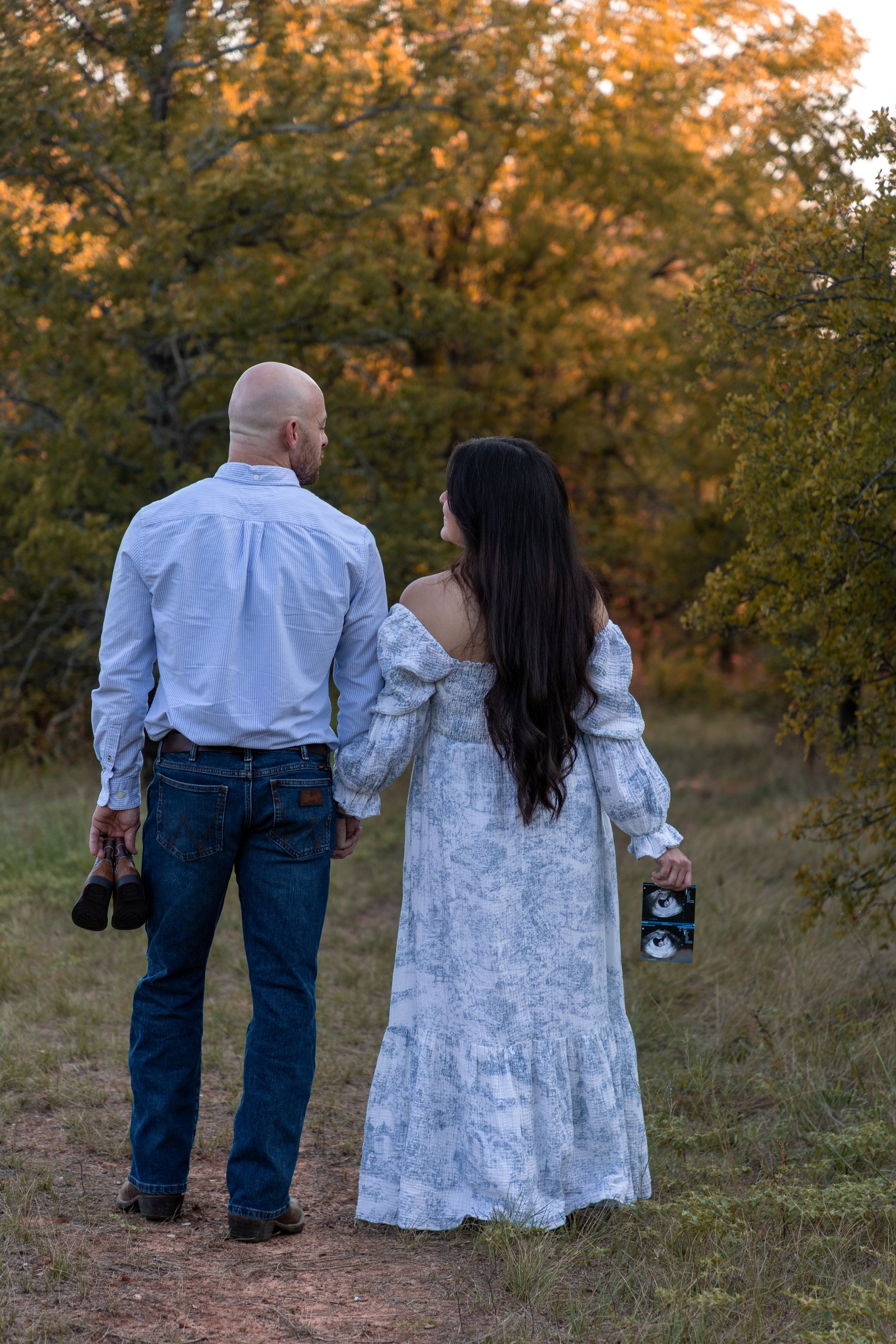 A couple holding hands walking through a wooded area during sunset; the woman is holding ultrasound images, and the man is holding binoculars.