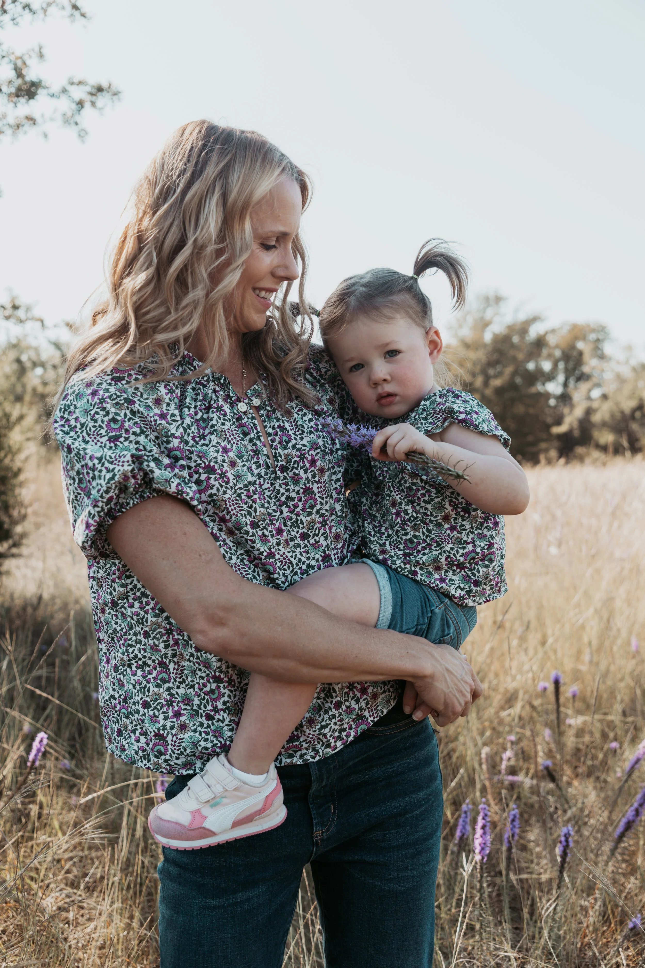 A woman with blonde curly hair holding a young girl in a field of tall grass and purple flowers on a sunny day.