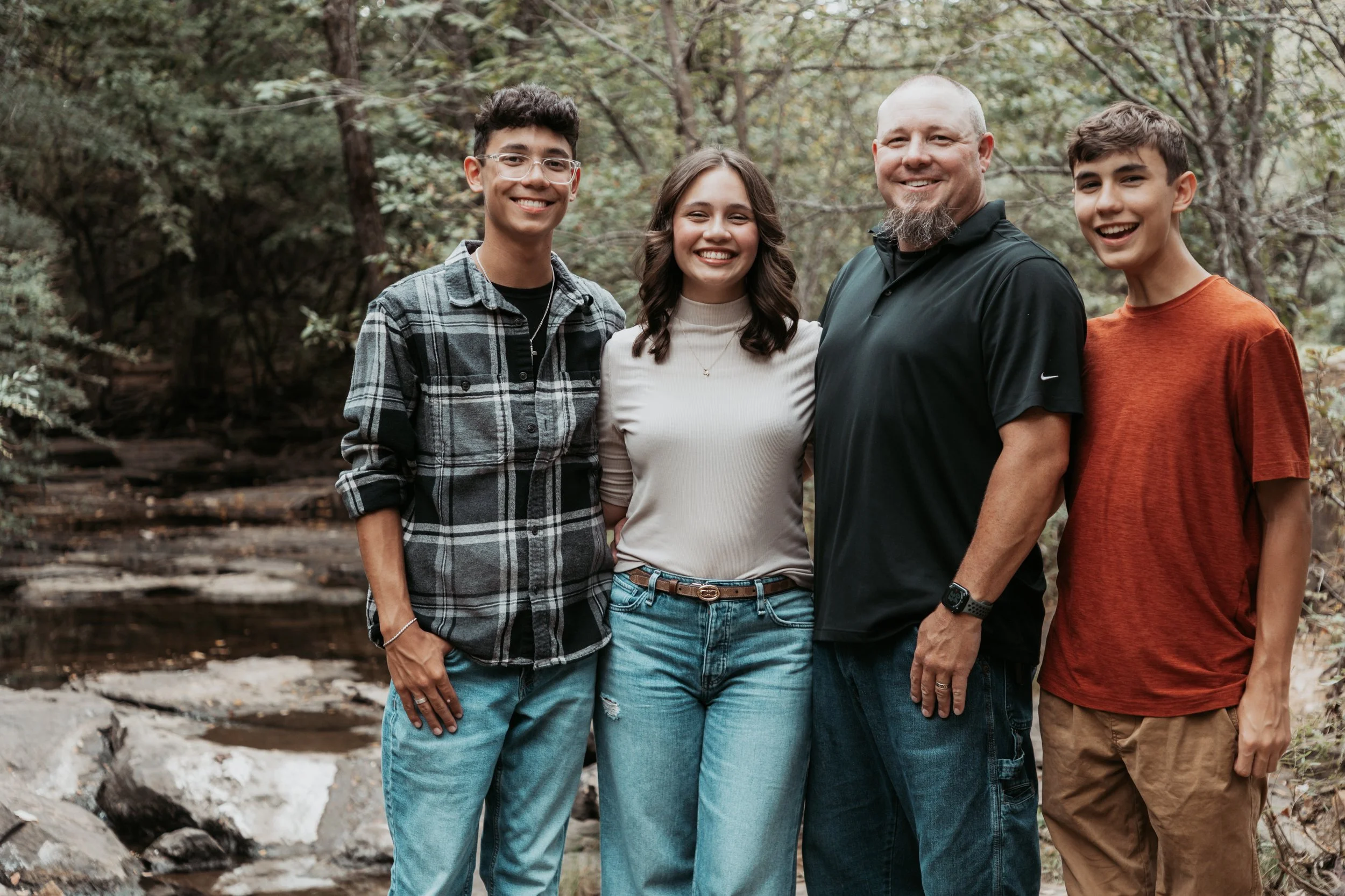A group of five people, including a man, a woman, and three teenagers, standing outdoors in a wooded area with a small creek in the background, smiling at the camera.