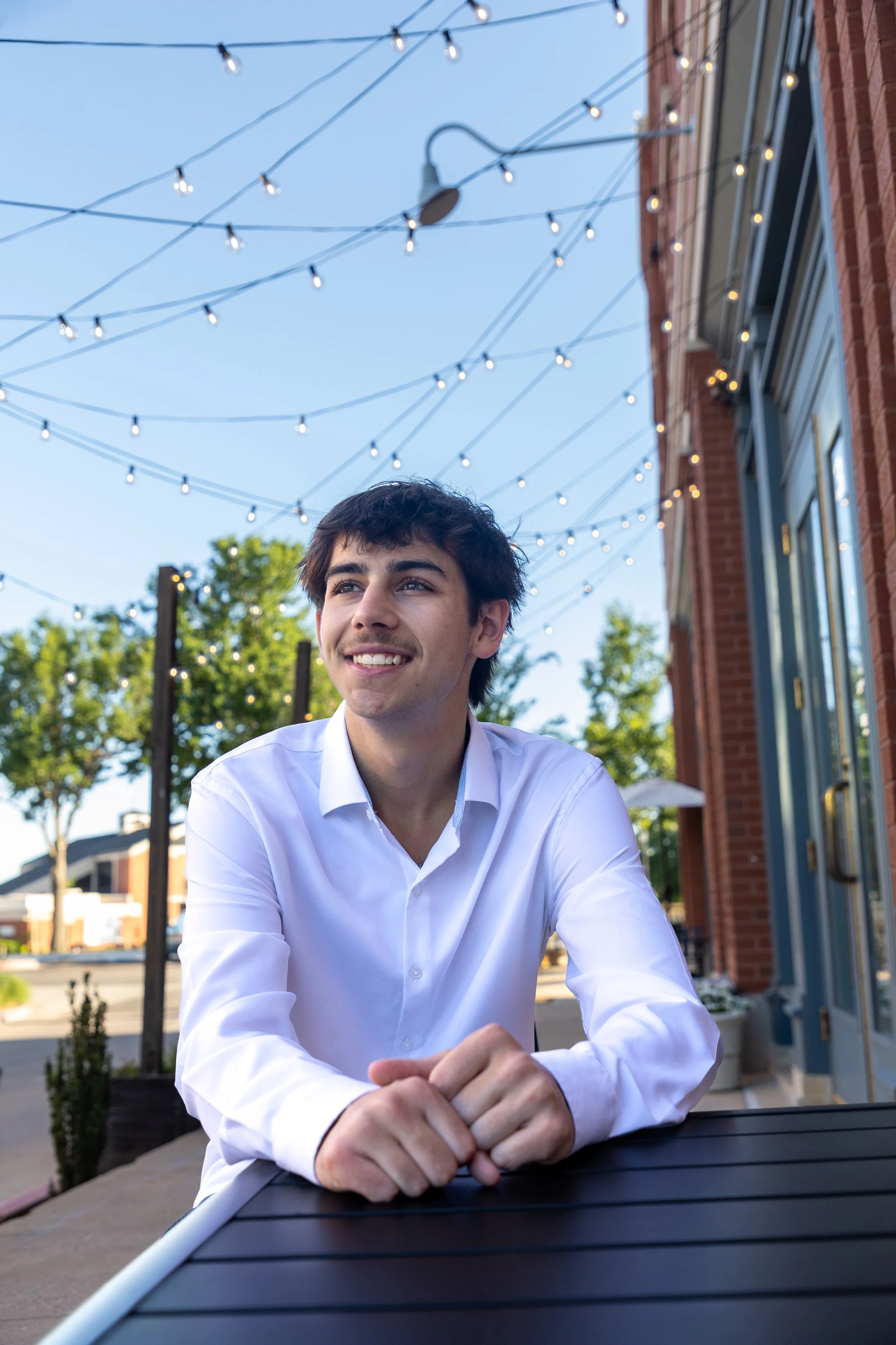 A young man with dark hair and a white shirt sitting outside at a table, smiling, with string lights overhead and trees and buildings in the background.