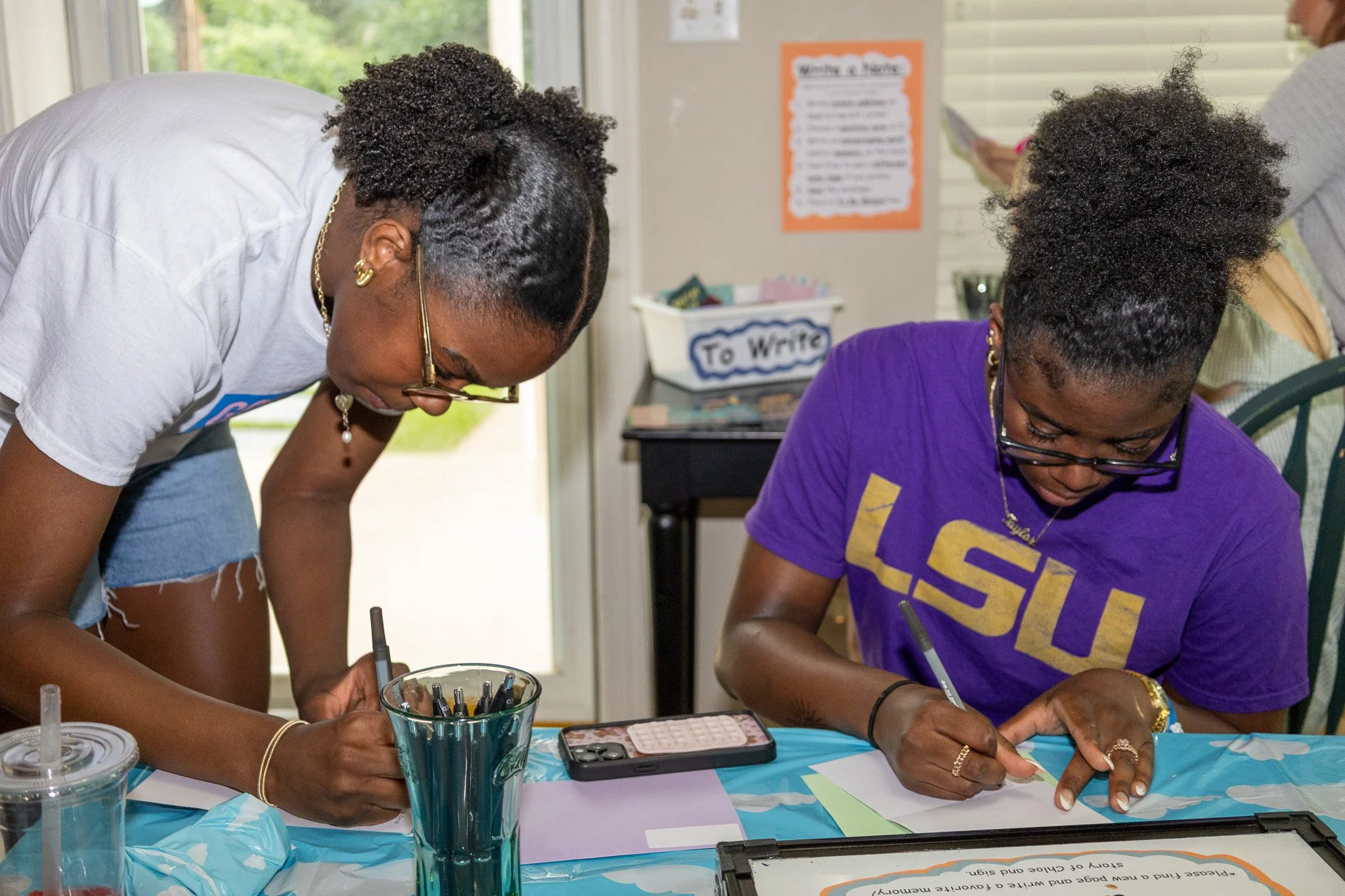 Two women with curly hair, wearing glasses, are writing at a table. One is in a white shirt and the other in a purple LSU t-shirt. The table has pens, paper, and a mobile phone. In the background, there are posters and a person partially visible to t