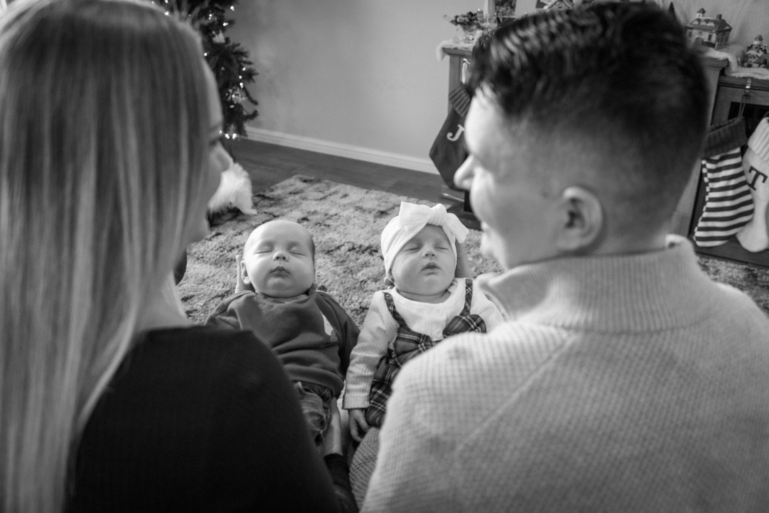 A family sitting on the floor, with two young children in the middle, surrounded by a Christmas tree and stockings in the background.