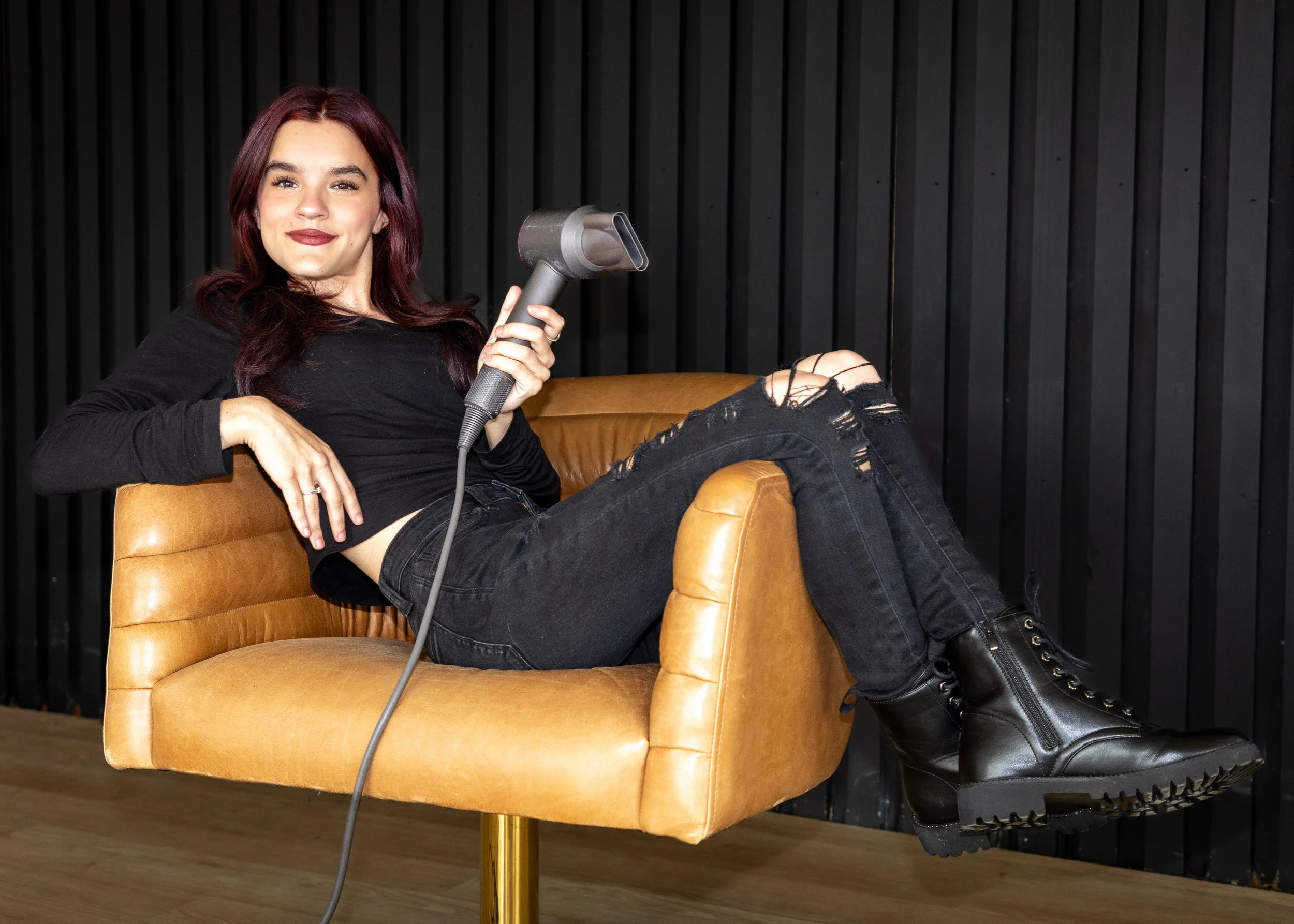 A woman with reddish hair sitting on a tan leather chair, holding a hairdryer, wearing a black long-sleeve top, ripped black jeans, and black combat boots, in front of a black wooden wall.