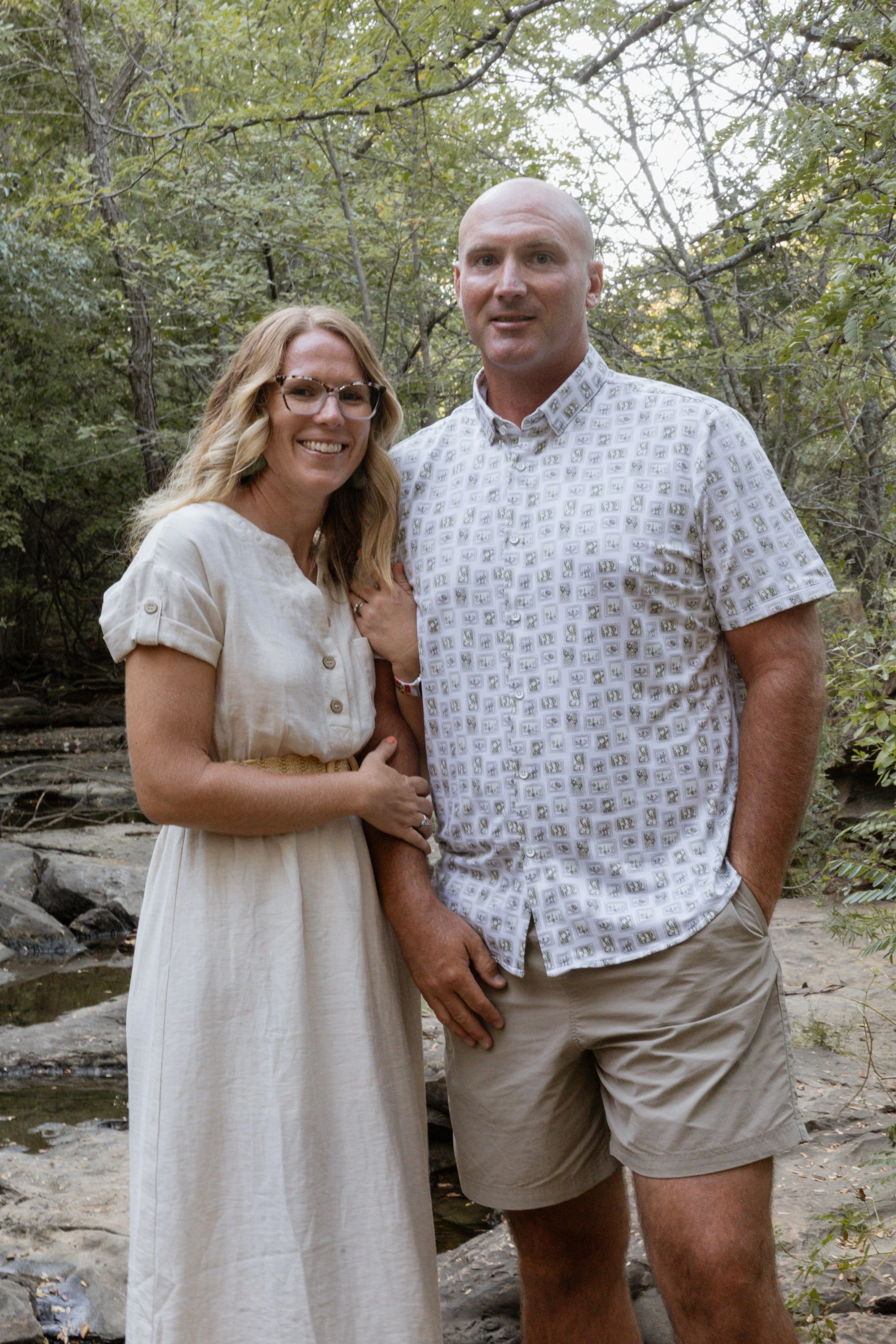 A smiling couple standing outdoors in a wooded area by a creek, the woman with blonde hair and glasses, the man with a shaved head, both dressed in light-colored summer clothing.