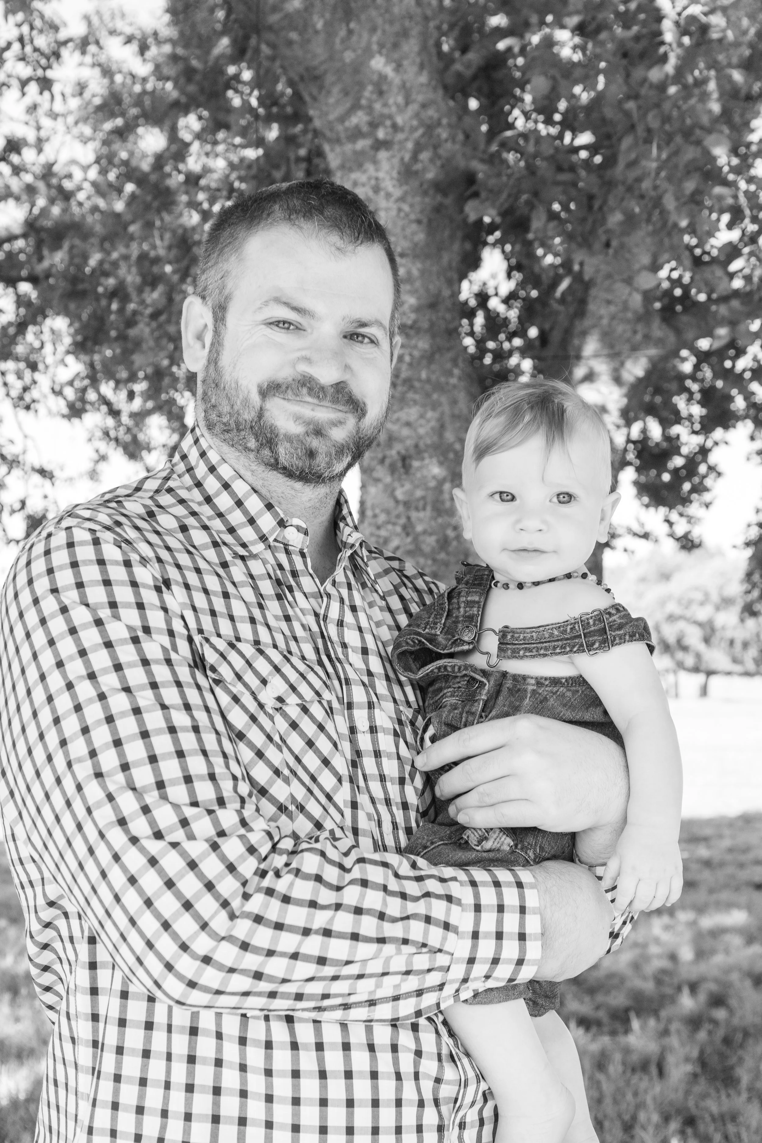 A man holding a young girl outdoors near a tree, both smiling, in black and white.