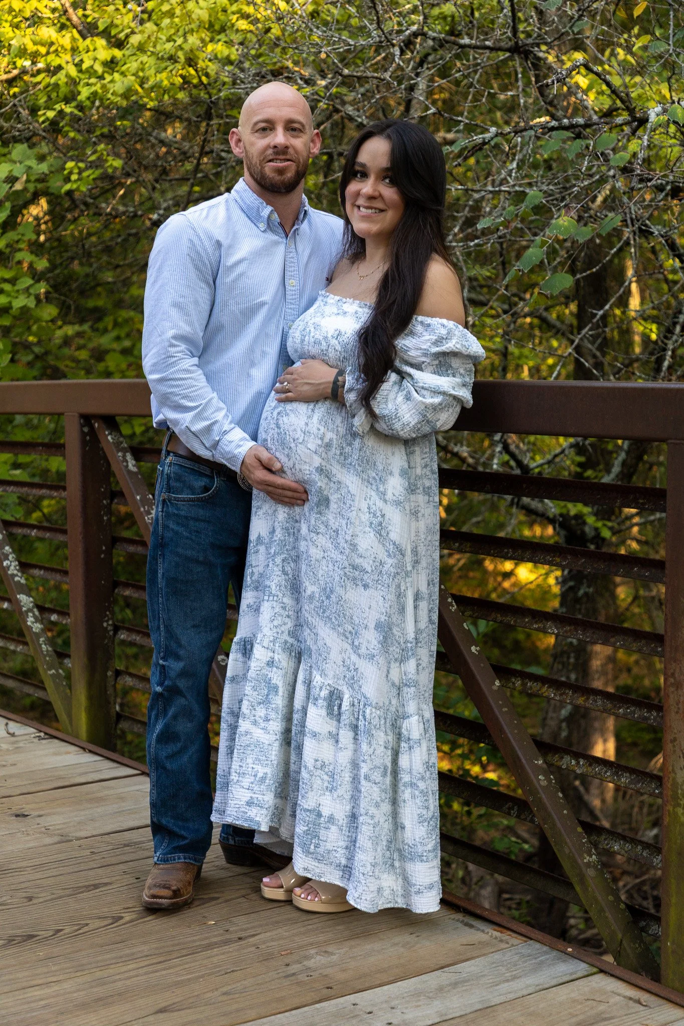 A couple standing on a wooden bridge in a wooded area, with trees and foliage in the background.