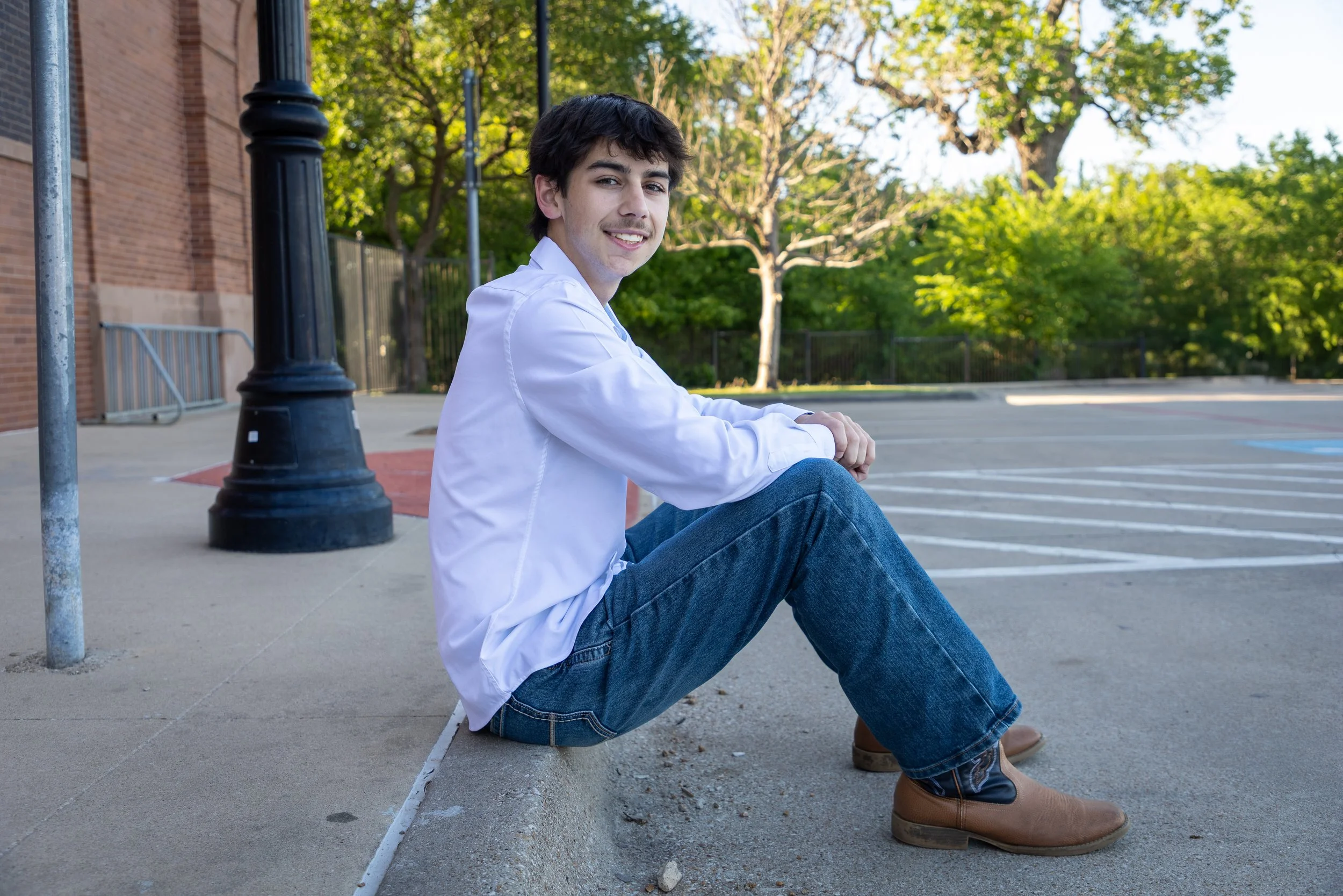 A young man sitting on a curb on a sidewalk, smiling at the camera, wearing a white shirt, blue jeans, and brown boots, with trees and a parking lot in the background.