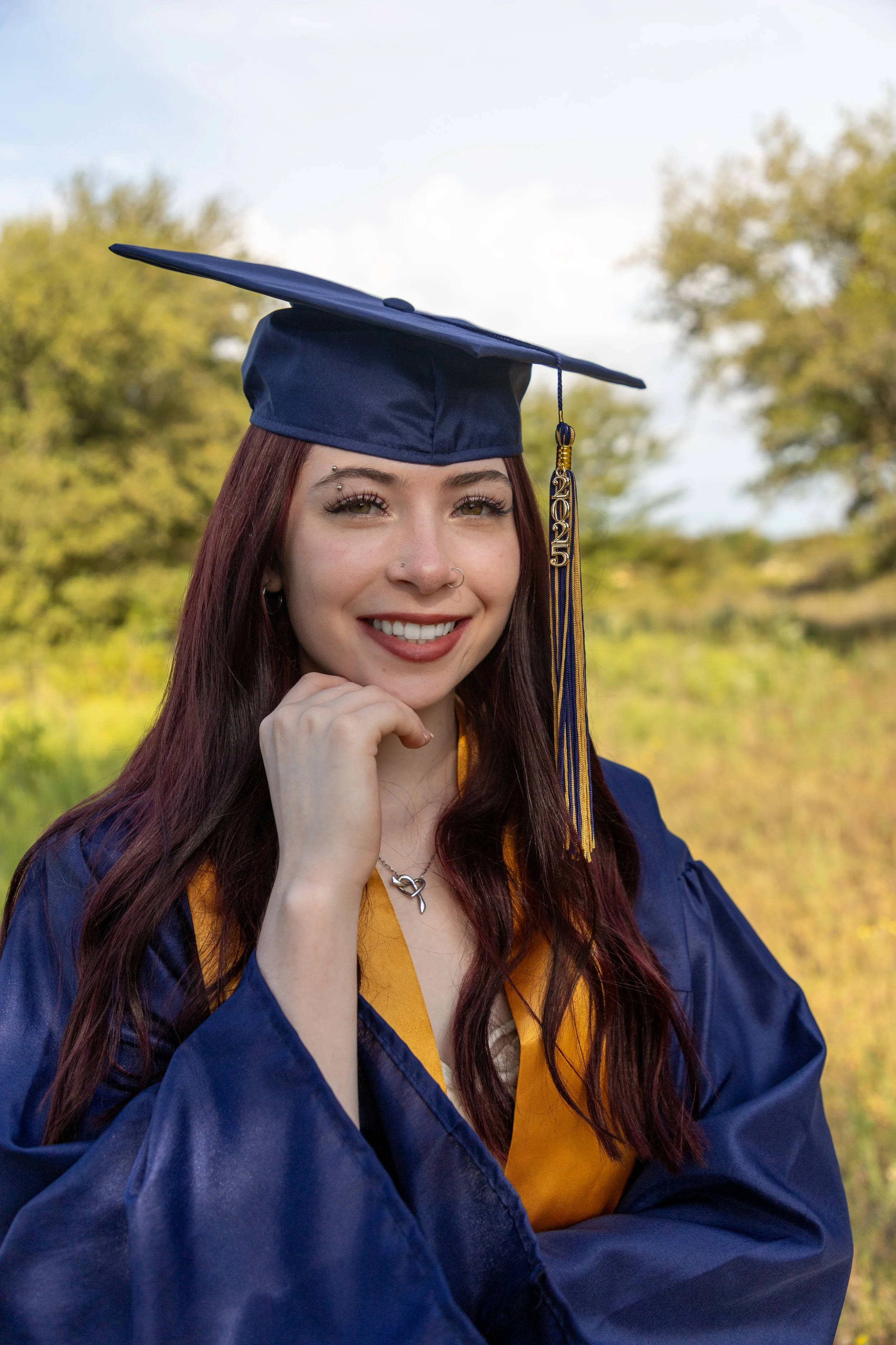 A young woman dressed in a blue graduation cap and gown, smiling outdoors with trees and a blue sky in the background.