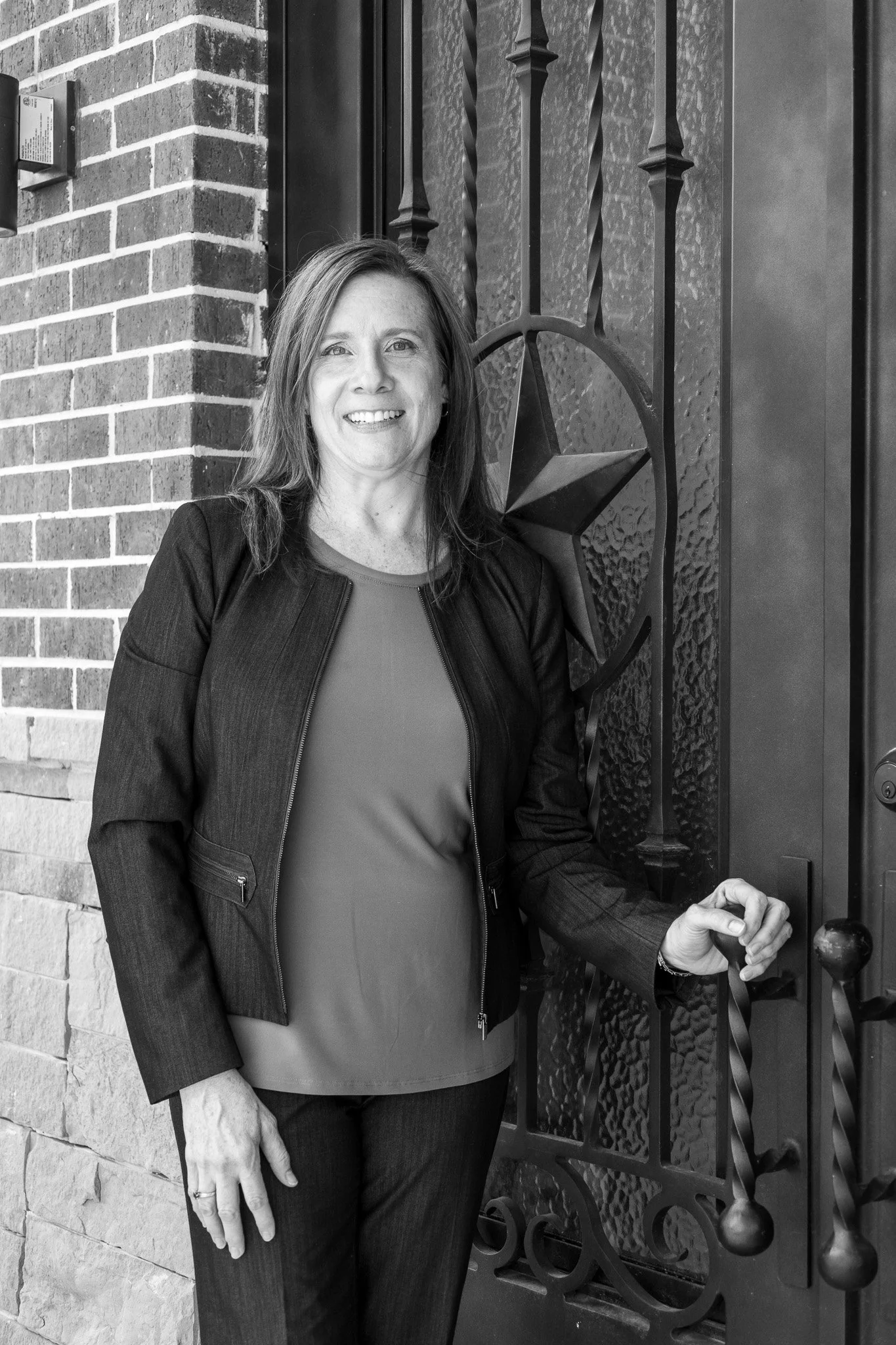 A woman with shoulder-length hair smiling, standing next to a decorative wrought iron gate with a star design, against a brick wall background.