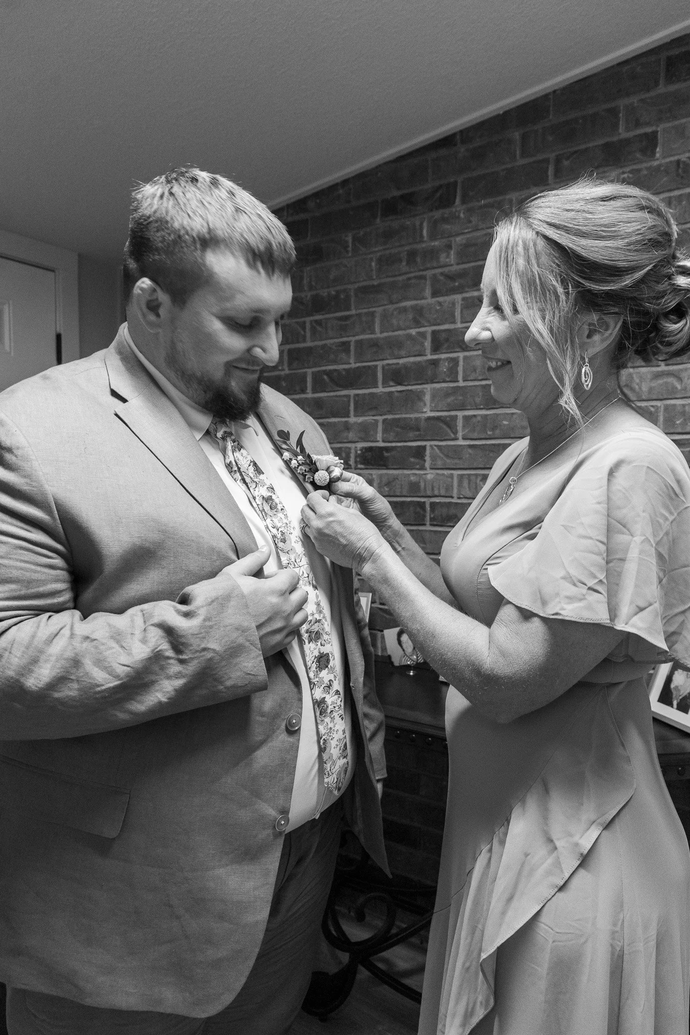 A woman pinning a boutonniere onto a man dressed in a suit, both smiling during a special occasion. The setting has a brick wall background.
