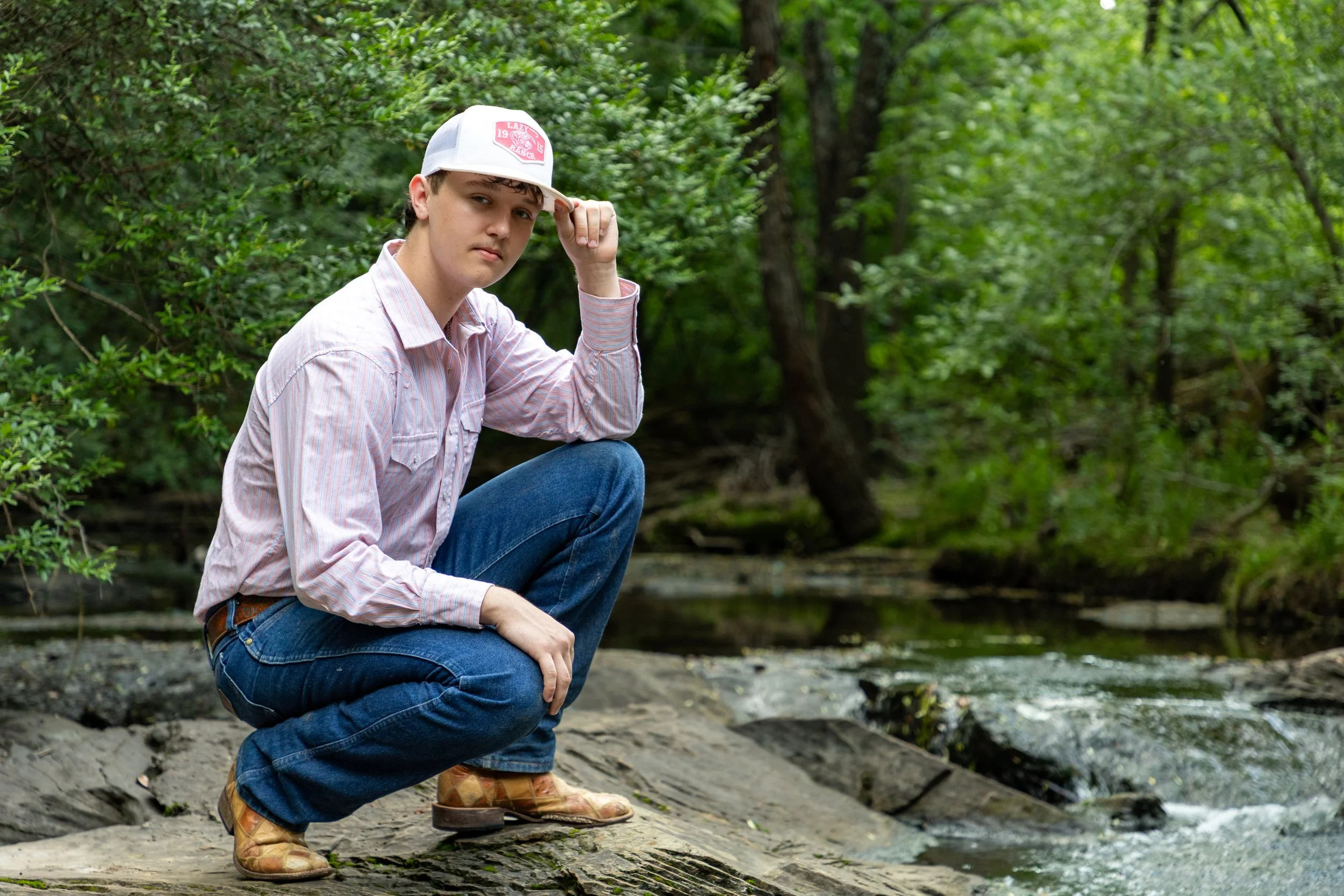 Young man in plaid shirt and jeans crouching by a creek in a wooded area, wearing a white baseball cap and looking at the camera.