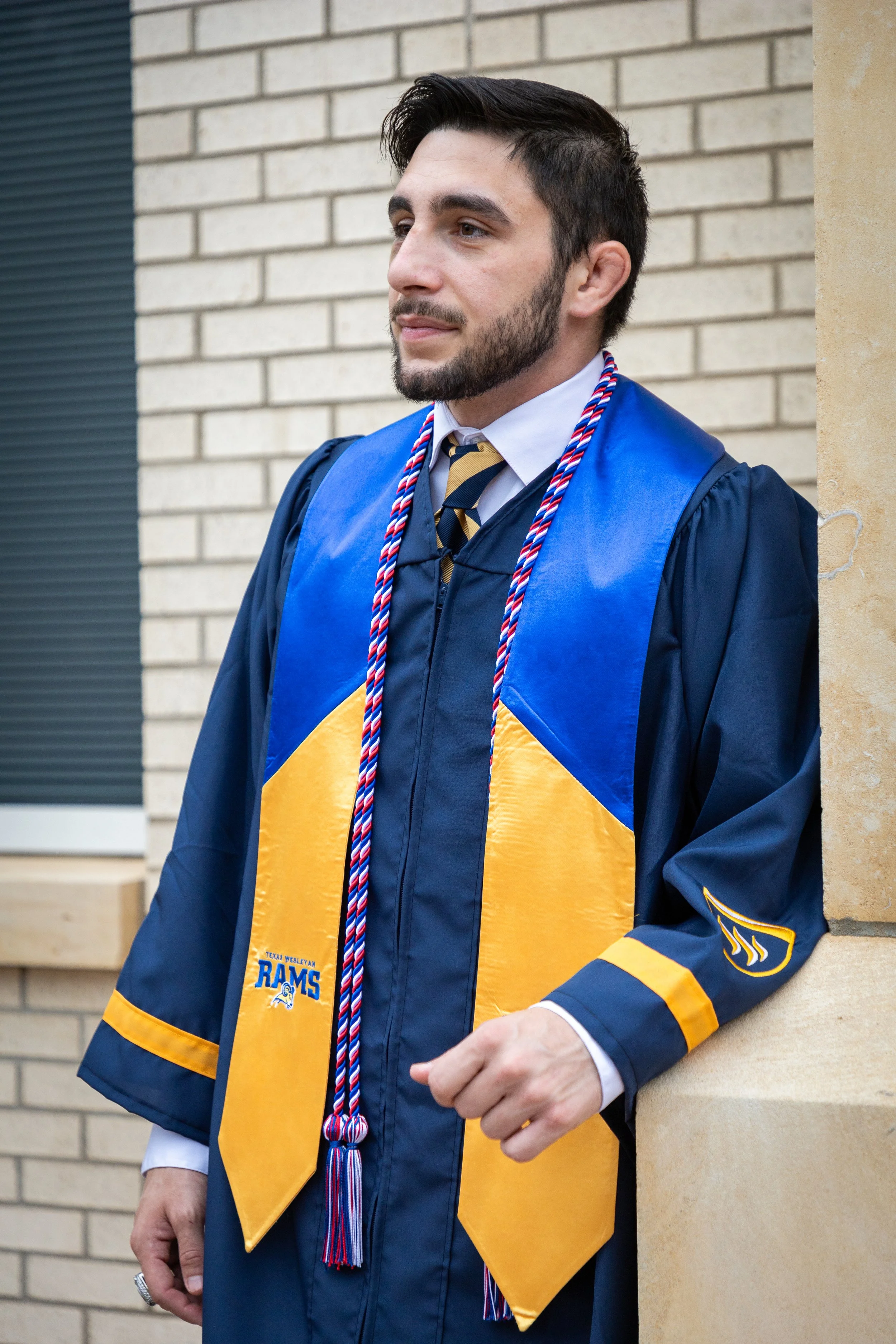 Young man in graduation cap and gown with cords, standing outdoors near a brick building.
