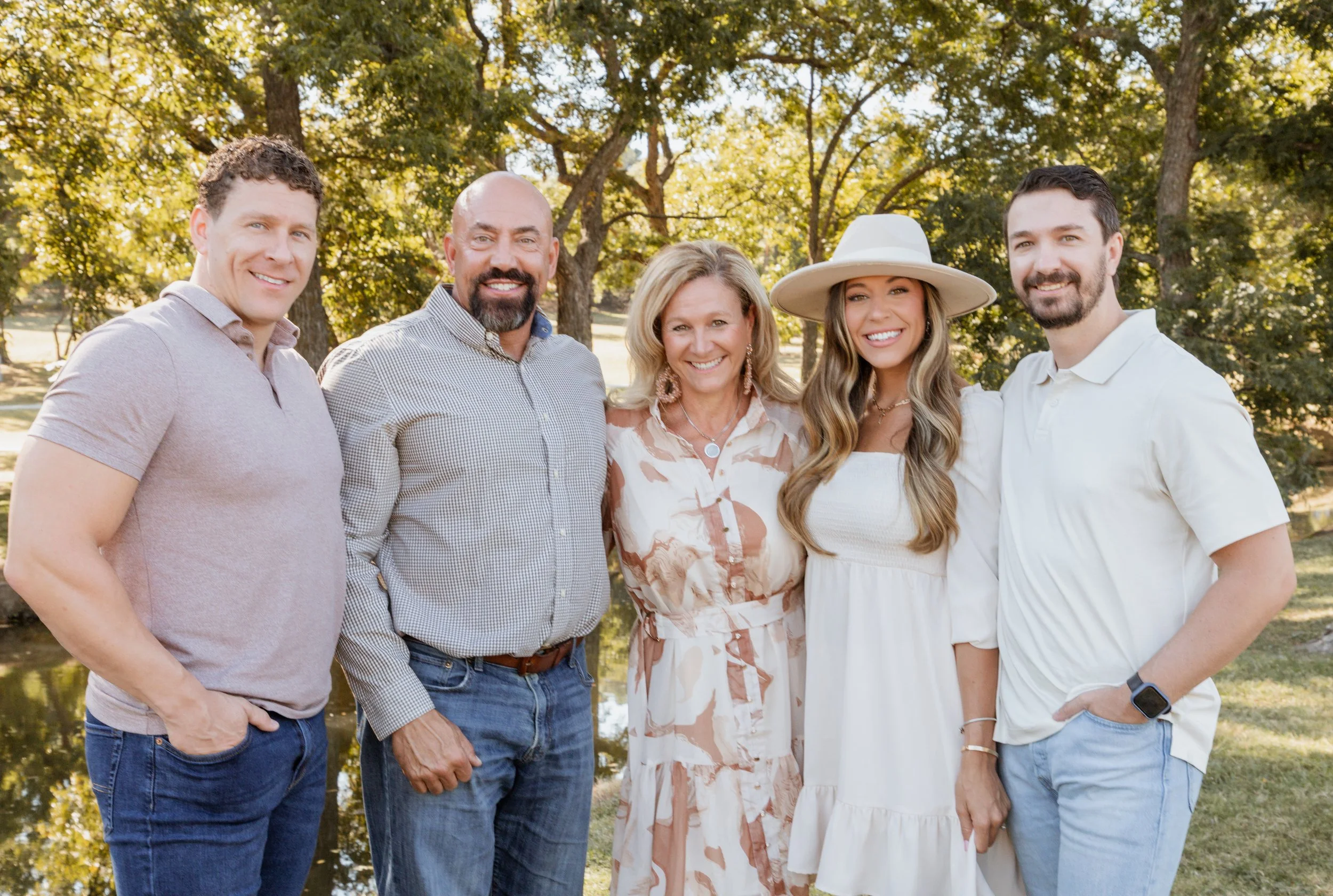 Five people standing outdoors by a pond, smiling and posing for a photo, with trees and sunlight in the background.