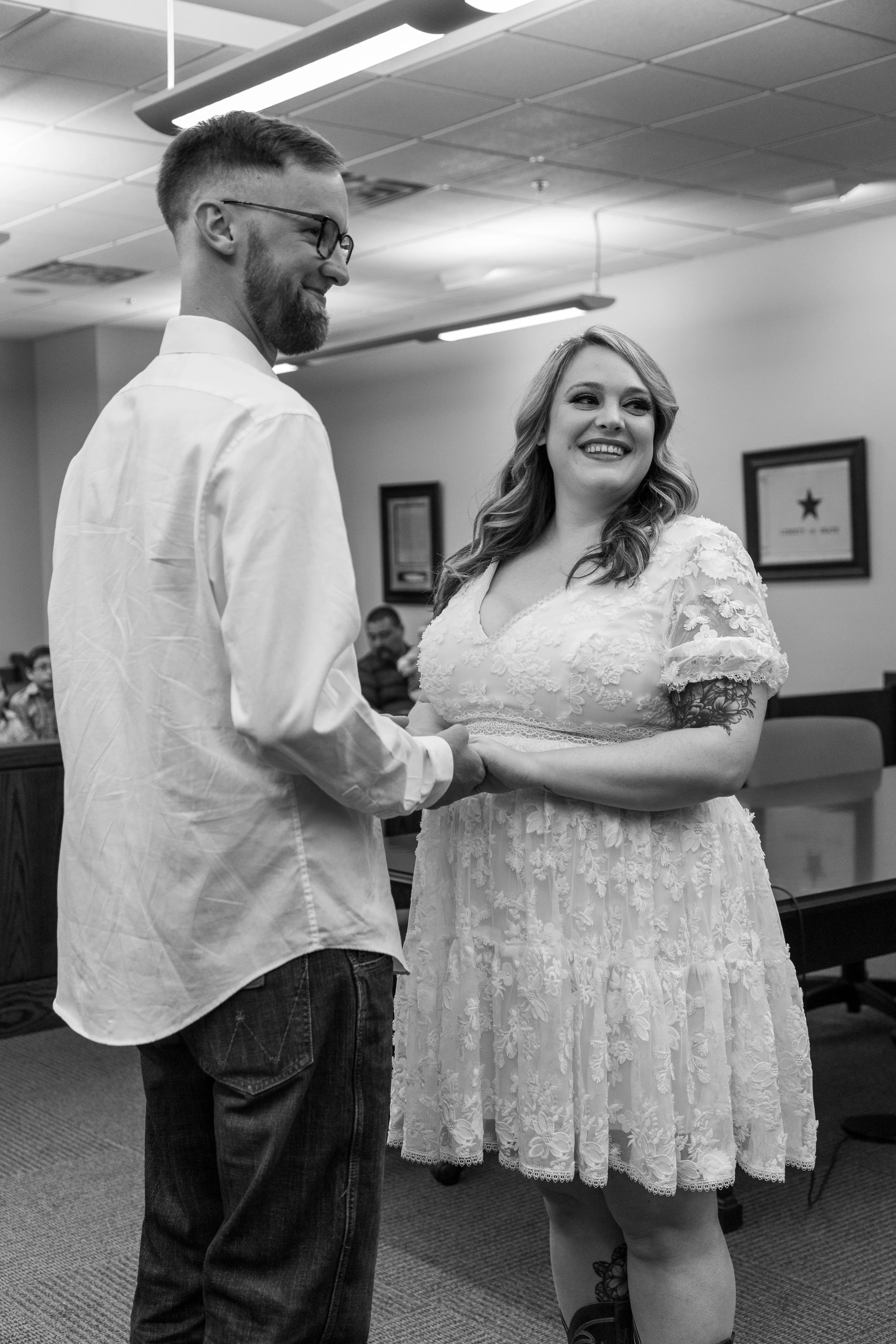 A black and white photo of a couple holding hands and smiling at each other during a wedding ceremony inside a room with framed certificates on the wall.