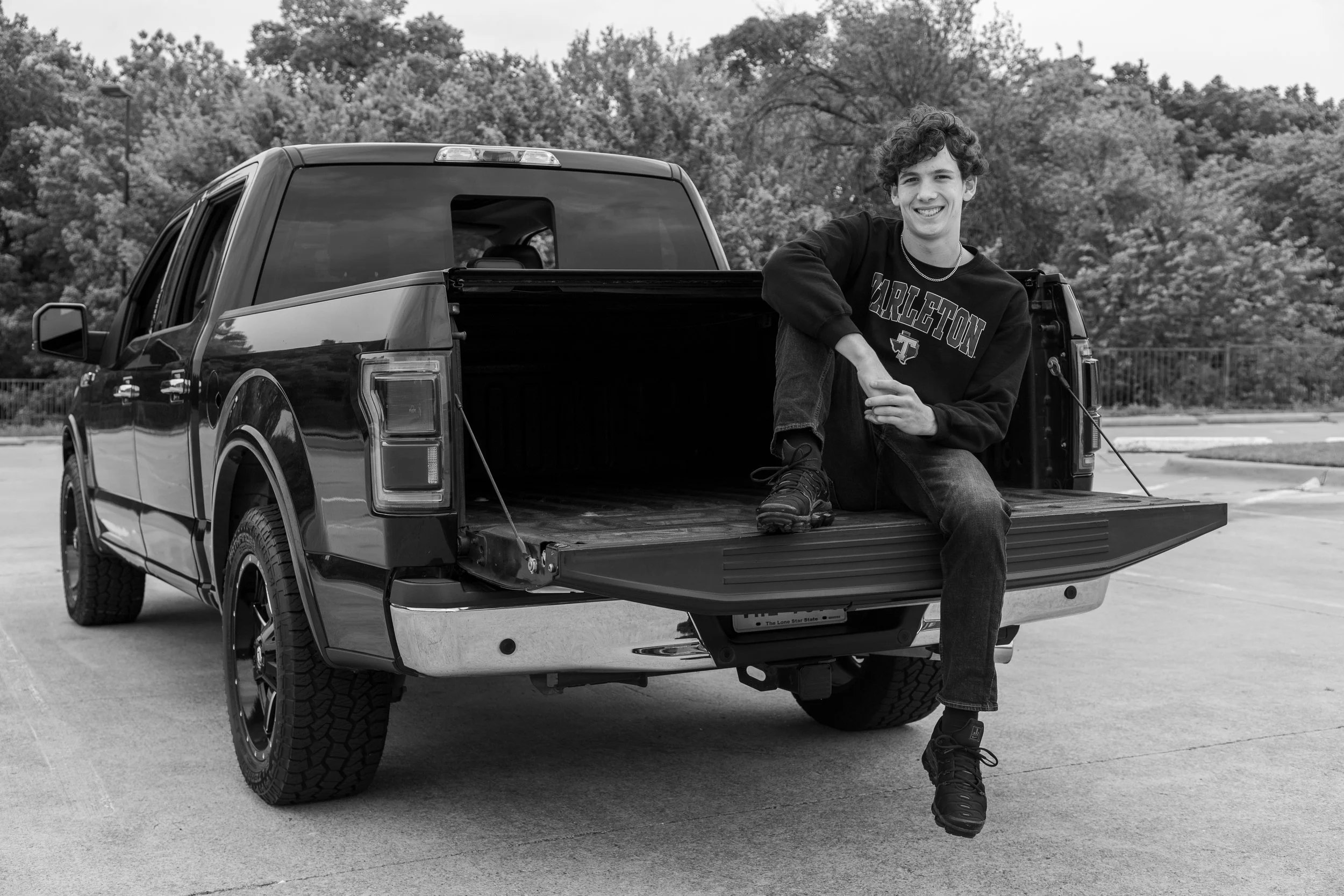 A young man sitting on the tailgate of a pickup truck in an outdoor parking lot, smiling at the camera. He is wearing a sweatshirt with a sports logo, jeans, and sneakers, with trees in the background.