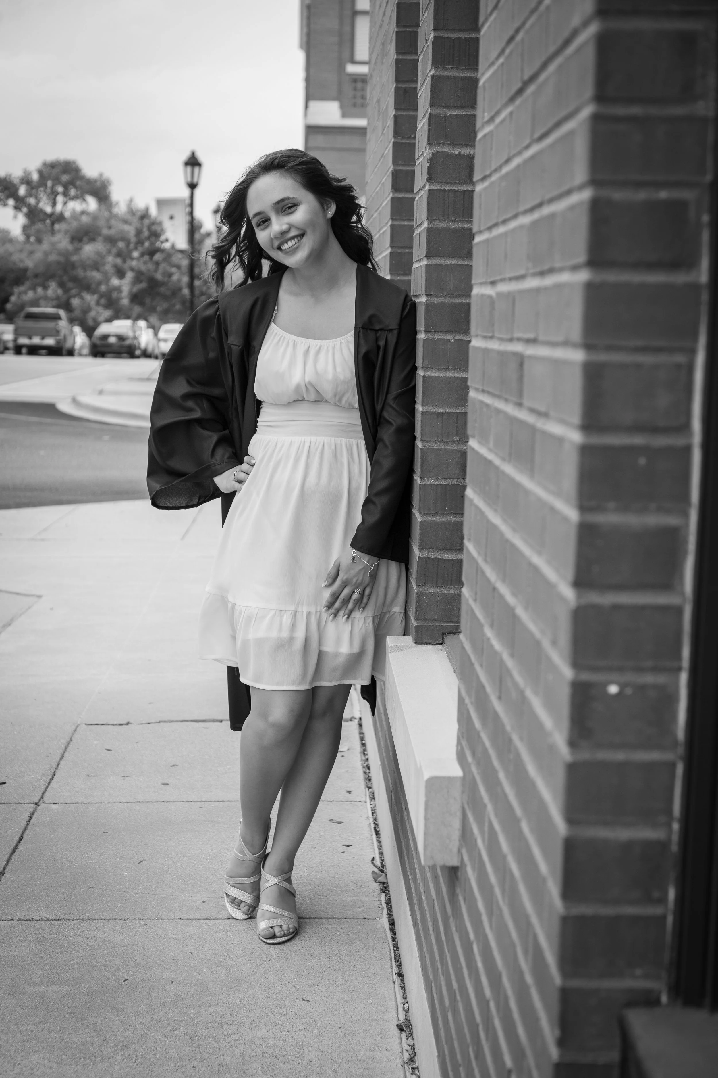 A young woman in a white dress and black jacket leaning against a brick wall, smiling outdoors.