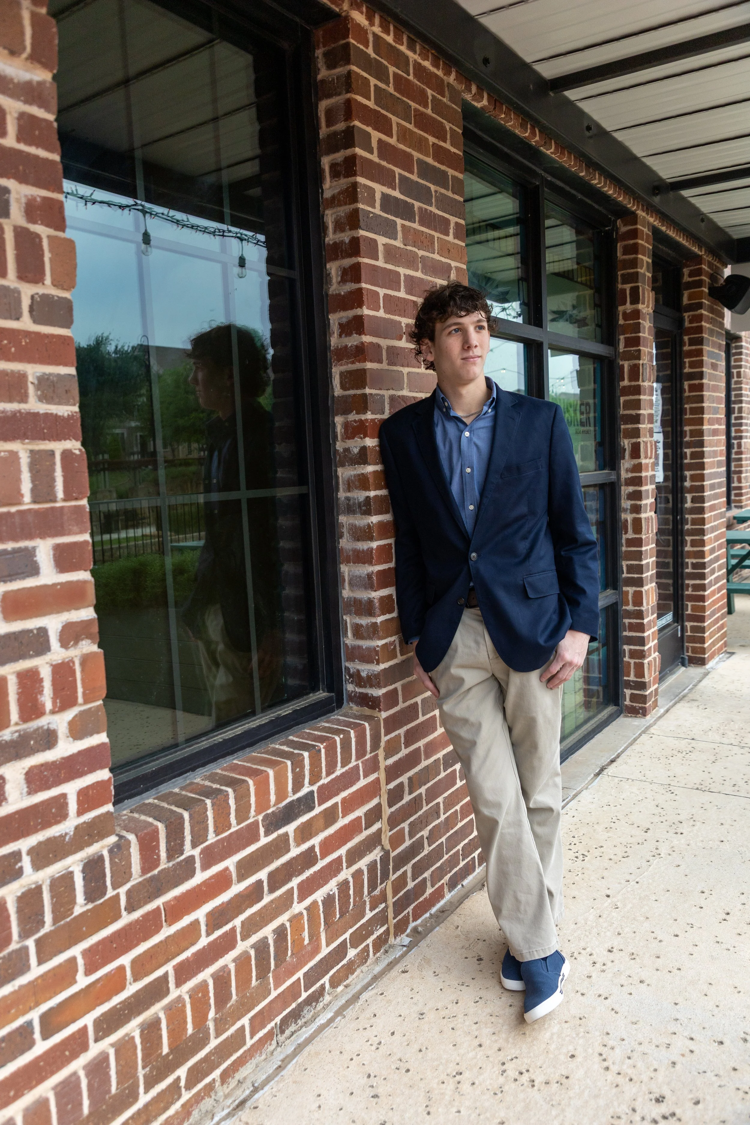 A young man in a navy blazer, blue shirt, khaki pants, and blue sneakers leaning against a brick wall outside a building with large glass windows.