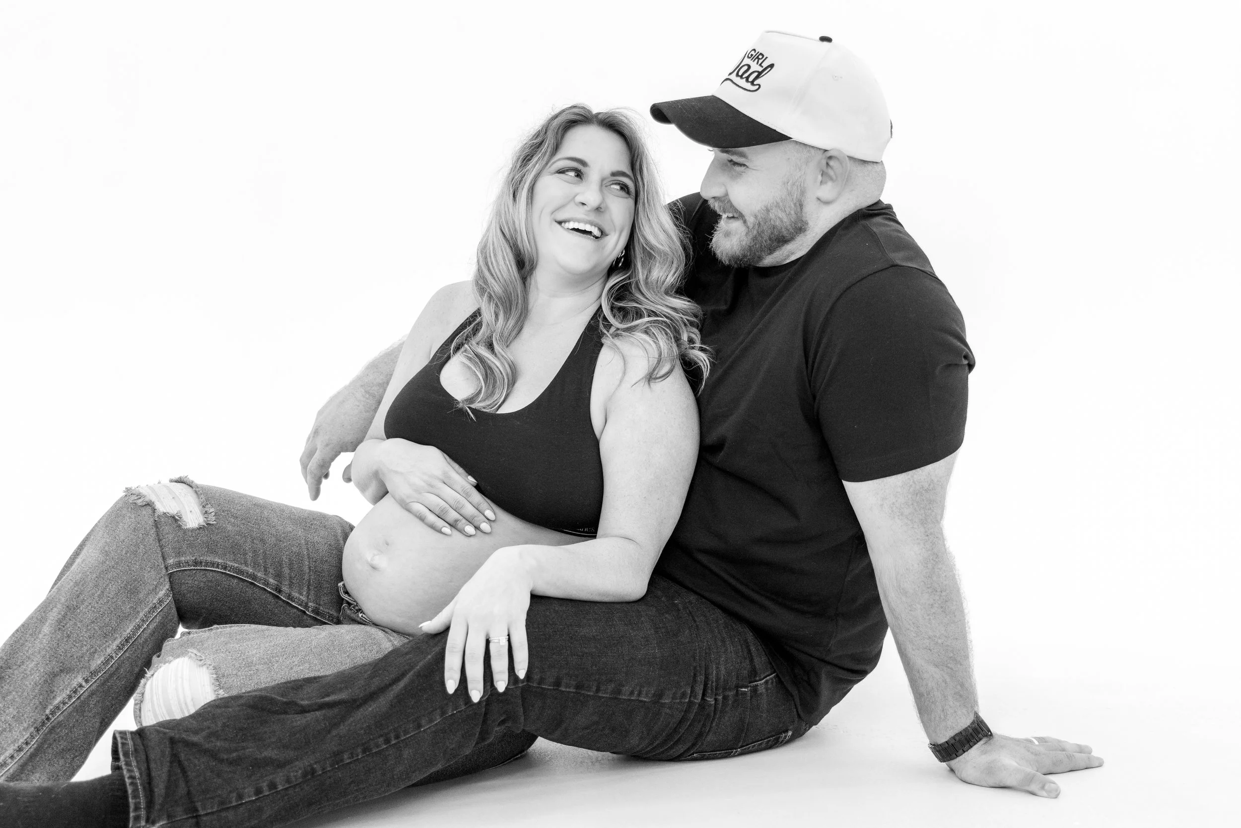A black and white photo of a smiling woman with long wavy hair and a man with a beard wearing a baseball cap. They are sitting close together, with the woman sitting on the man's lap, both looking at each other affectionately.