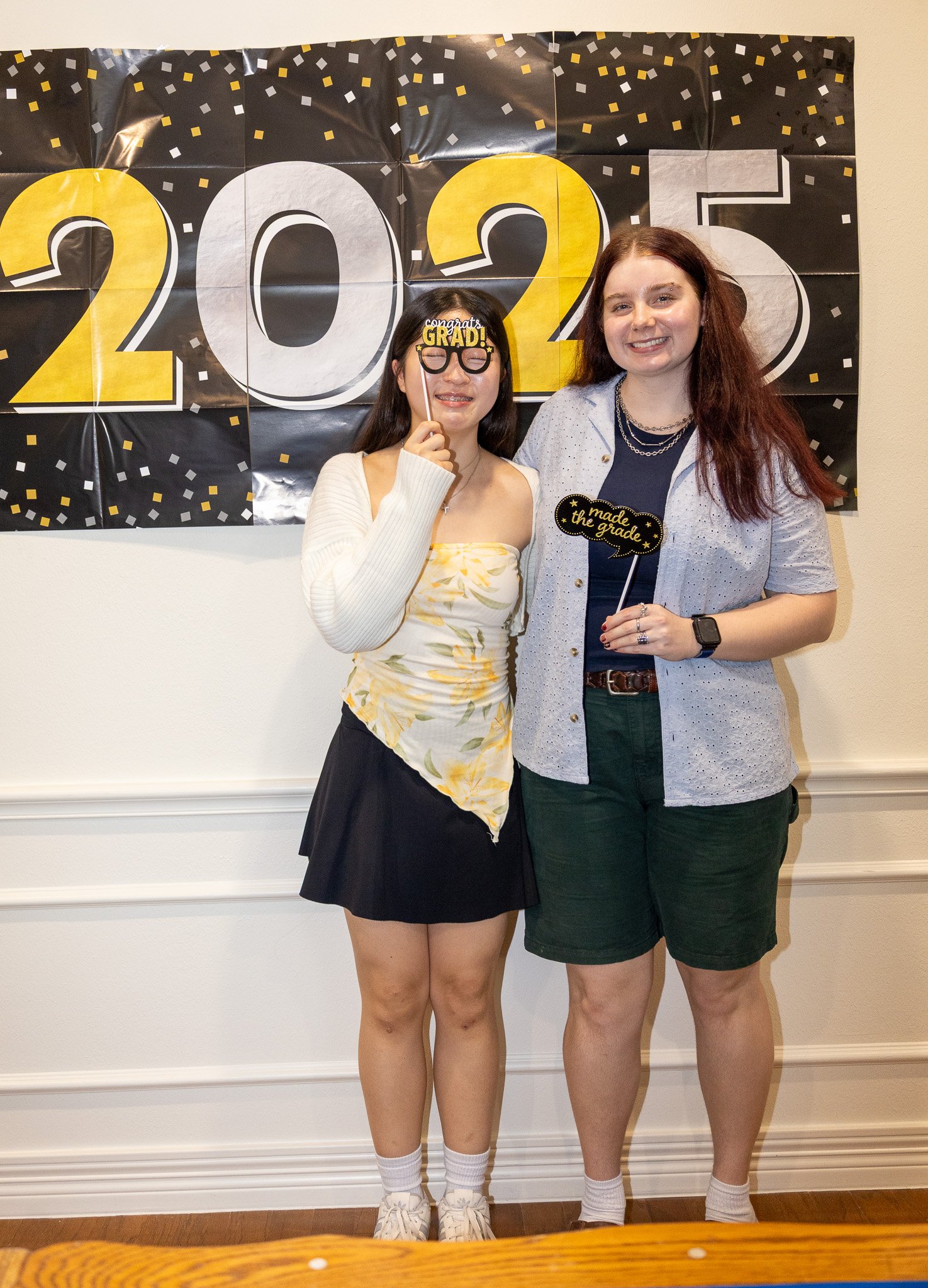 Two women standing in front of a black and yellow graduation celebration banner for 2023. One woman is holding a "congrats grad" sign and the other is holding a "made the grade" sign.
