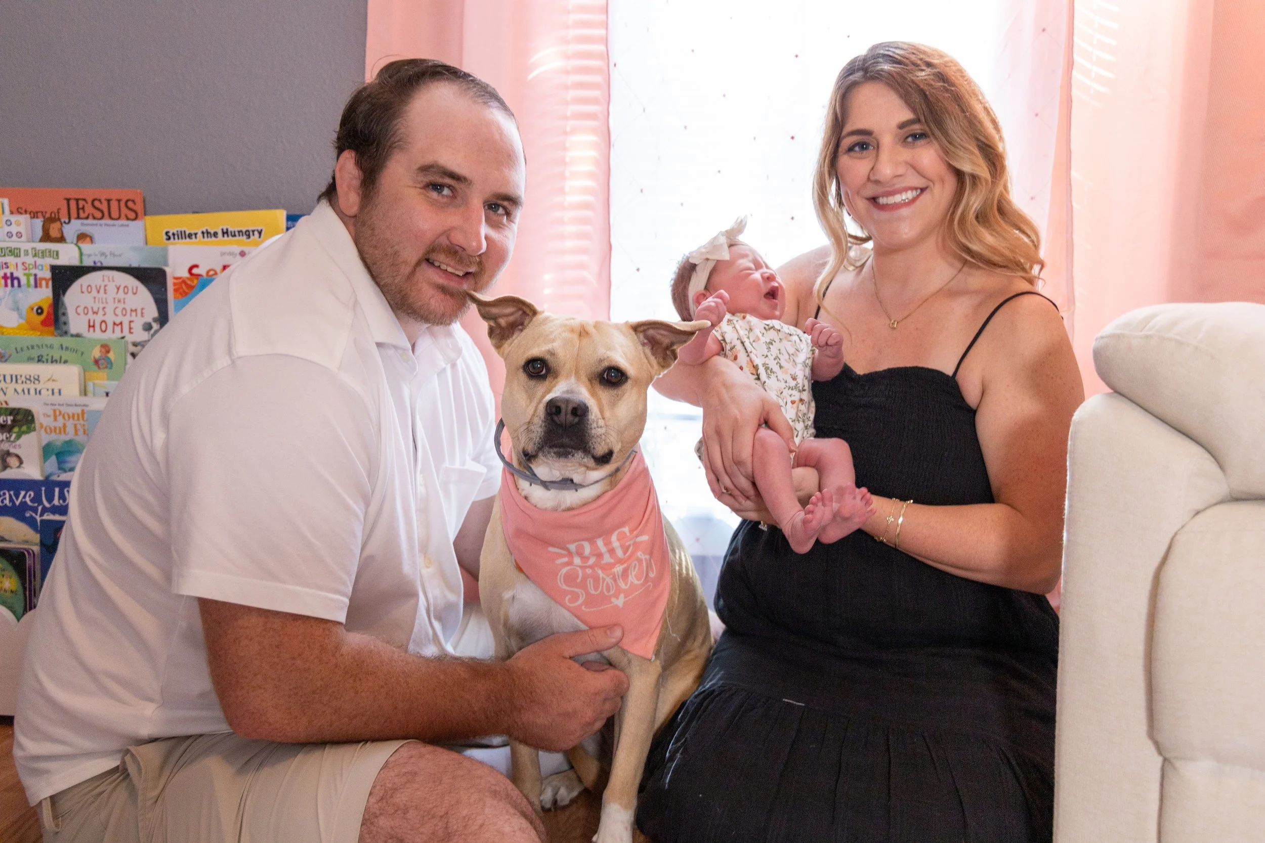 A man and woman sit close together holding a dog and a baby in a cozy room with bookshelves. The man has short brown hair and is wearing a white shirt. The woman has long, wavy hair and is wearing a black top. The dog, a tan and white breed, is weari