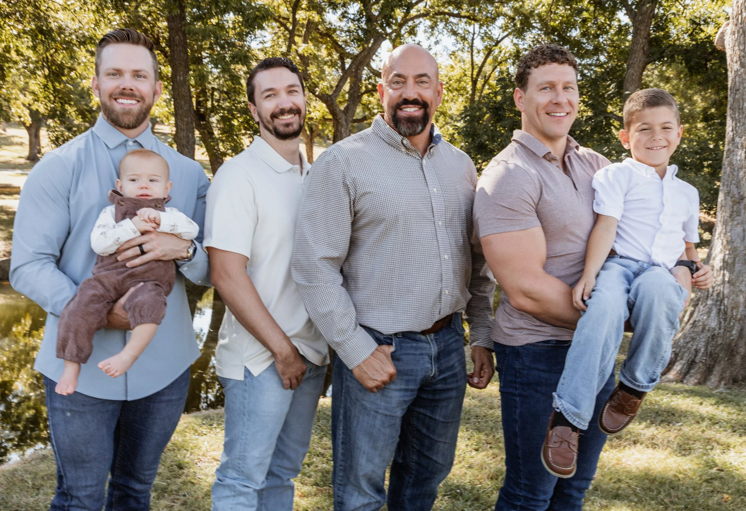 Group of six males, including children, outdoors in a park with trees and water in the background, smiling at the camera.