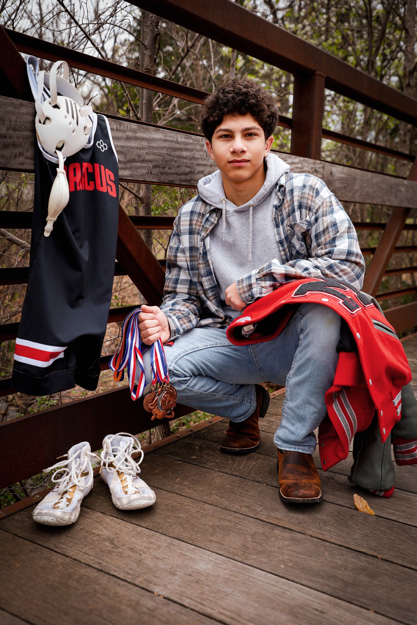 A teenage boy at an outdoor wooden bridge, dressed casually in a hoodie, jeans, and boots, kneeling next to a sports jacket with medals, a helmet, and a pair of white sneakers placed on the bridge floor.