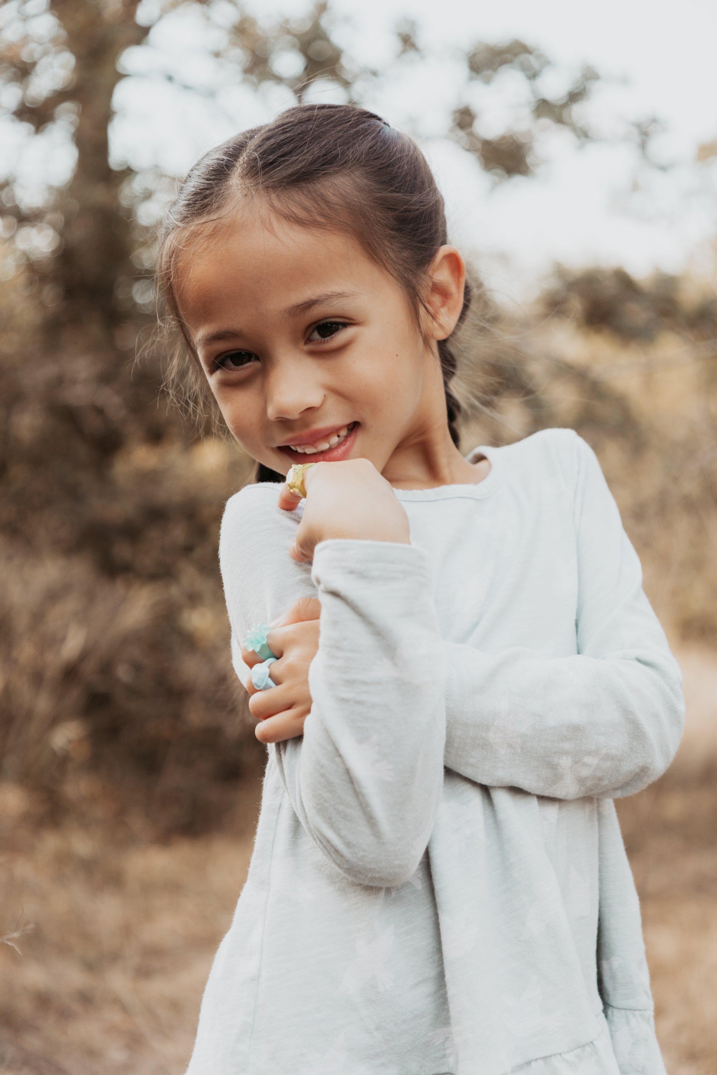 A young girl with dark hair in a braid smiling outdoors in a natural setting with trees and foliage in the background.