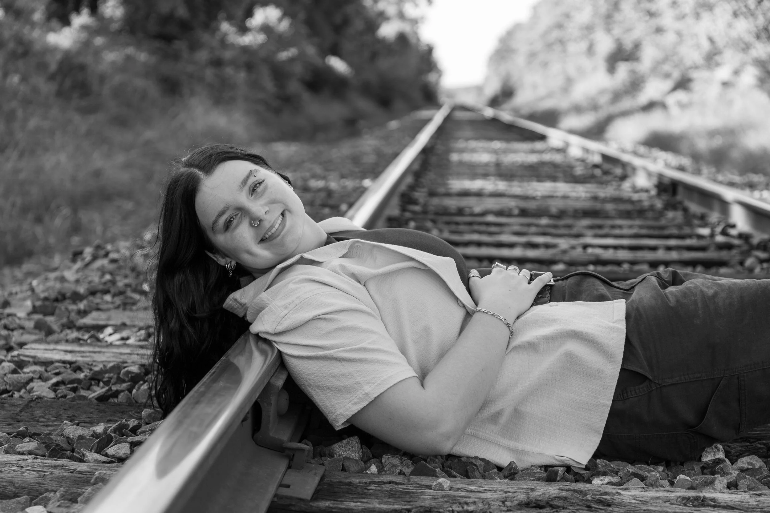 A young woman with dark hair and a septum piercing lying on her back on a railway track, smiling at the camera, in an outdoor setting with trees in the background.