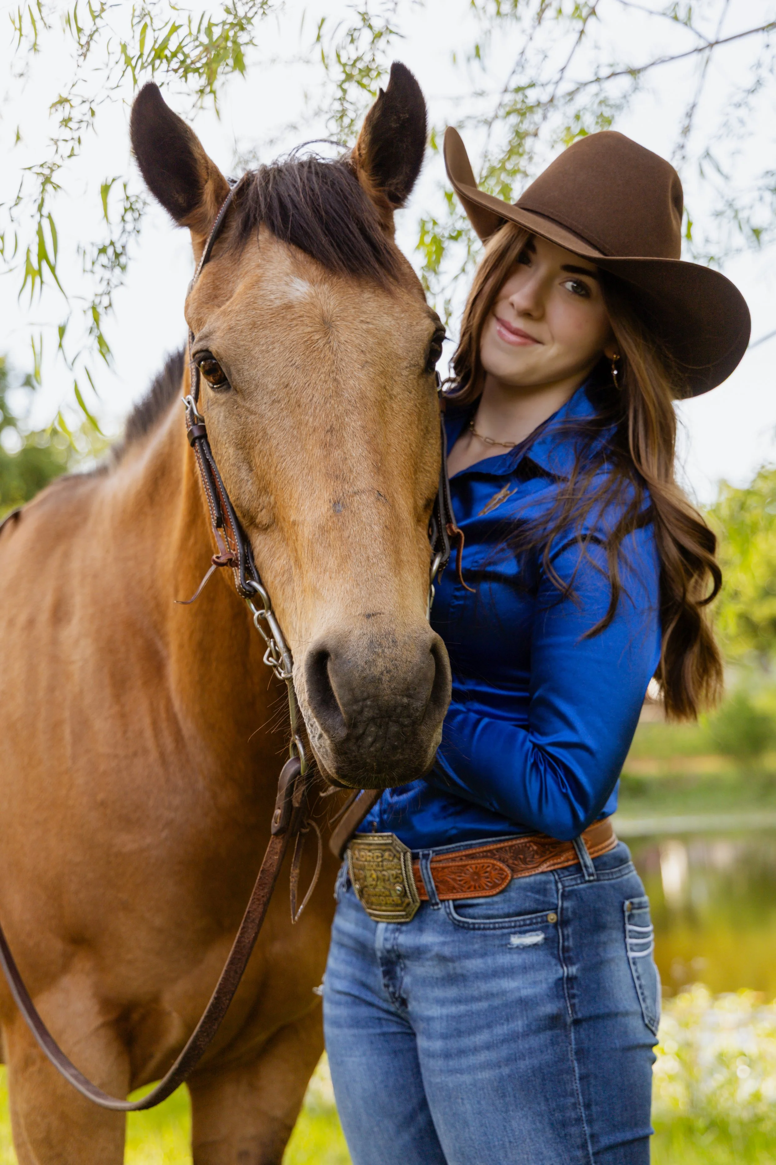 A young woman in a blue shirt and jeans standing next to a light brown horse with a dark mane. She is wearing a wide-brimmed brown hat and smiling. The background shows green leaves and a partly cloudy sky.