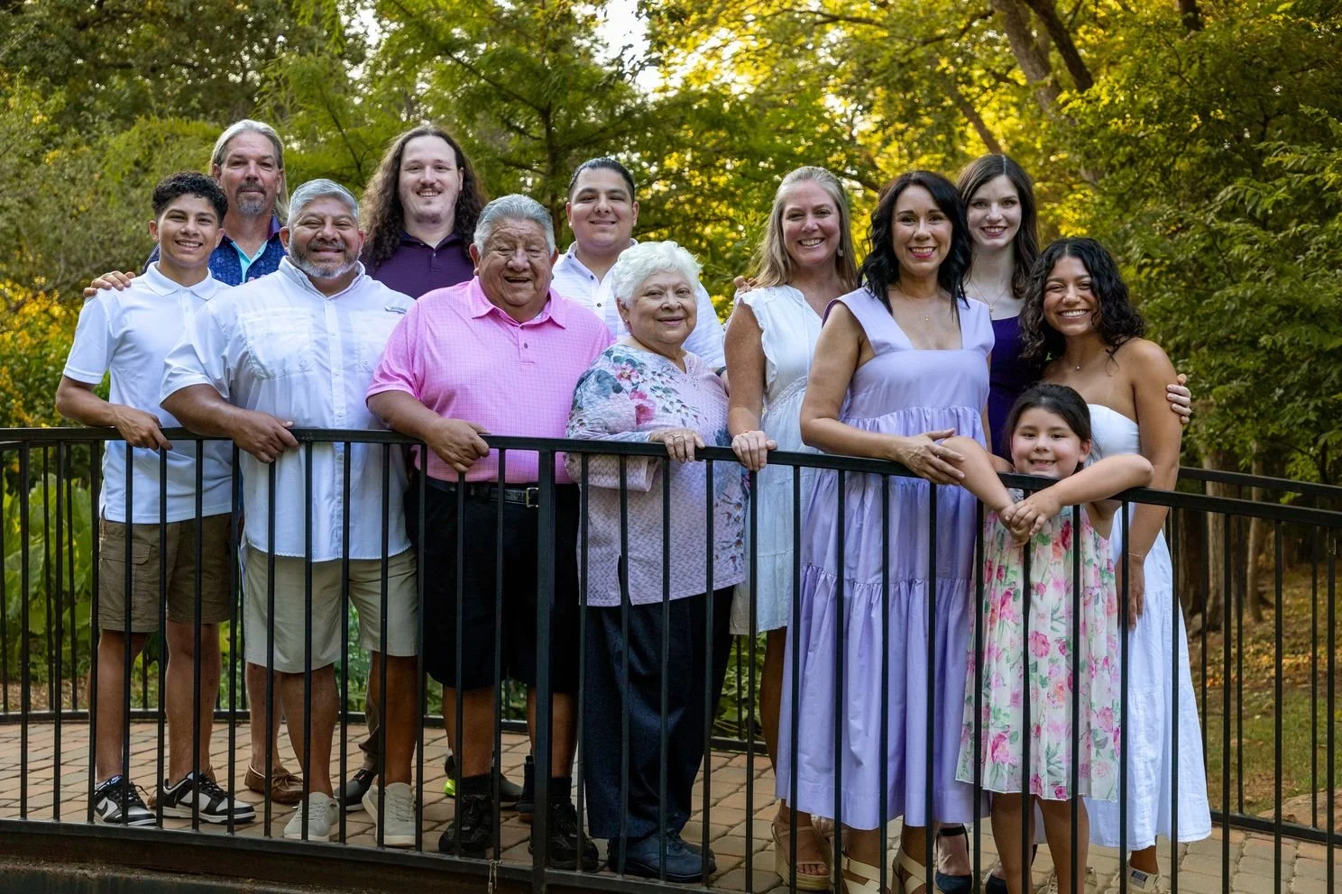 A multigenerational family of thirteen people standing on a stone patio behind a black metal railing outdoors among green trees, smiling at the camera.