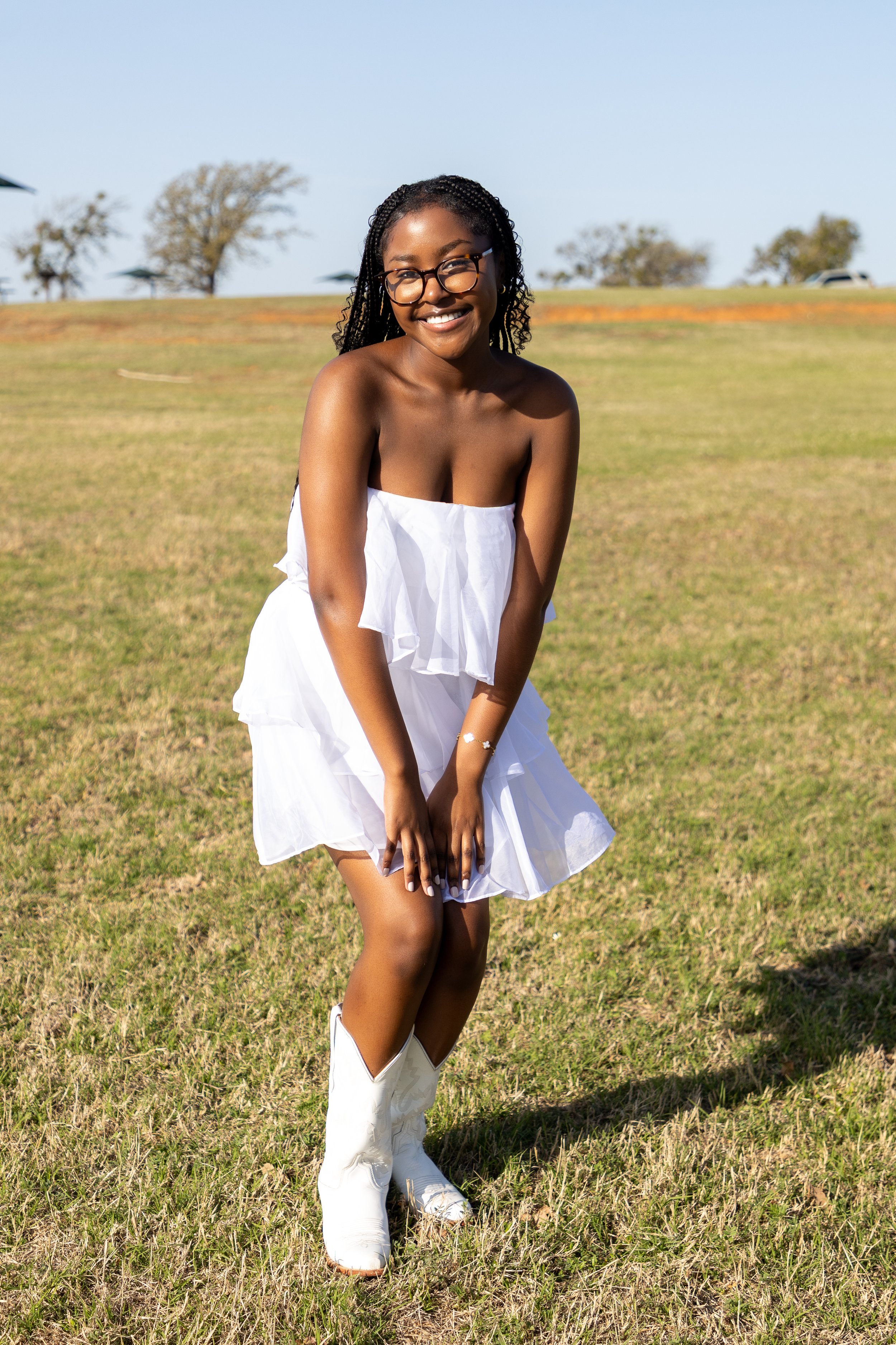 Young woman smiling outdoors in a white strapless dress and white cowboy boots, standing on grass with trees and a clear blue sky in the background.