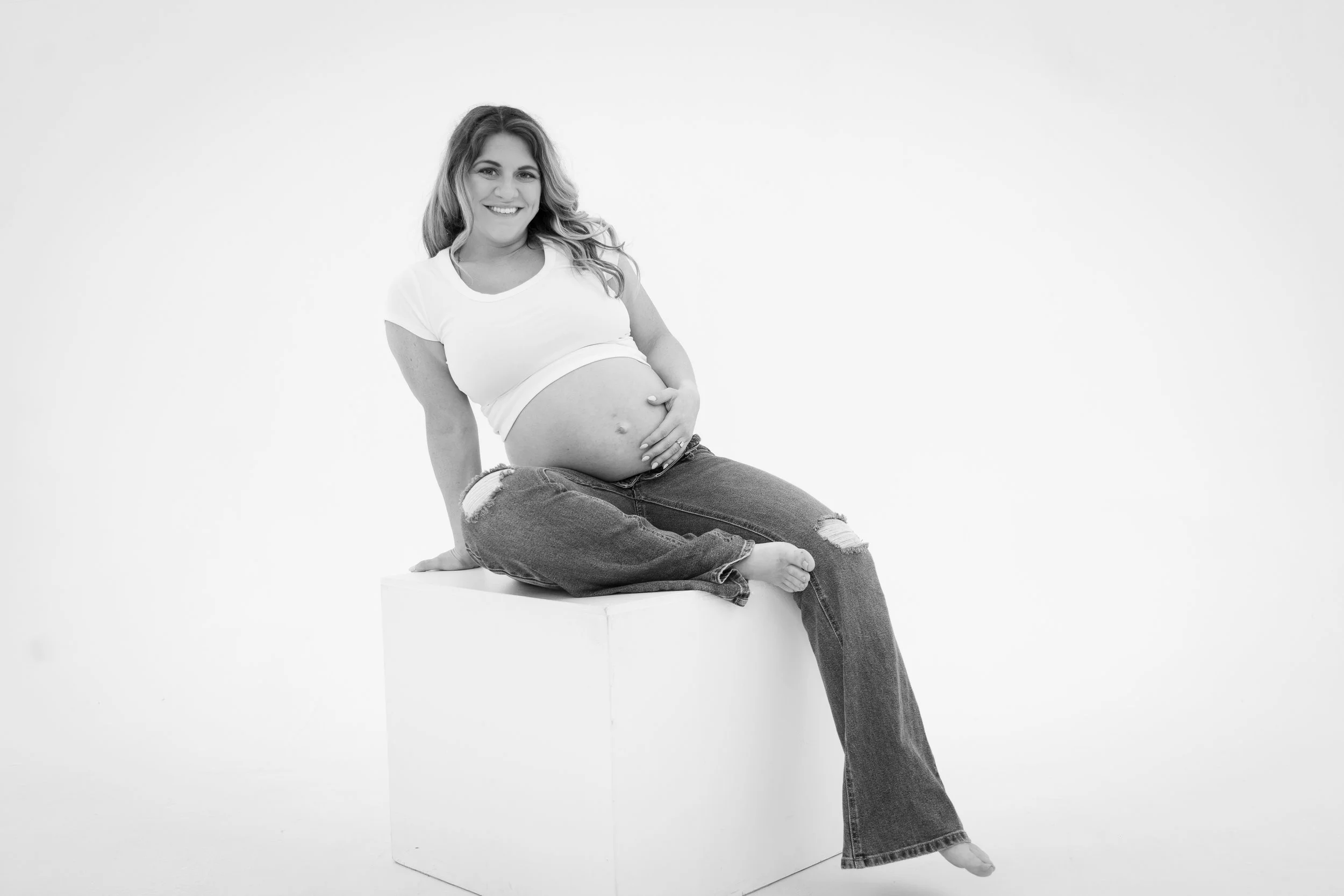 A pregnant woman with long, wavy hair wearing a white t-shirt and ripped jeans, sitting on a block against a plain white background, smiling and resting her hand on her belly.